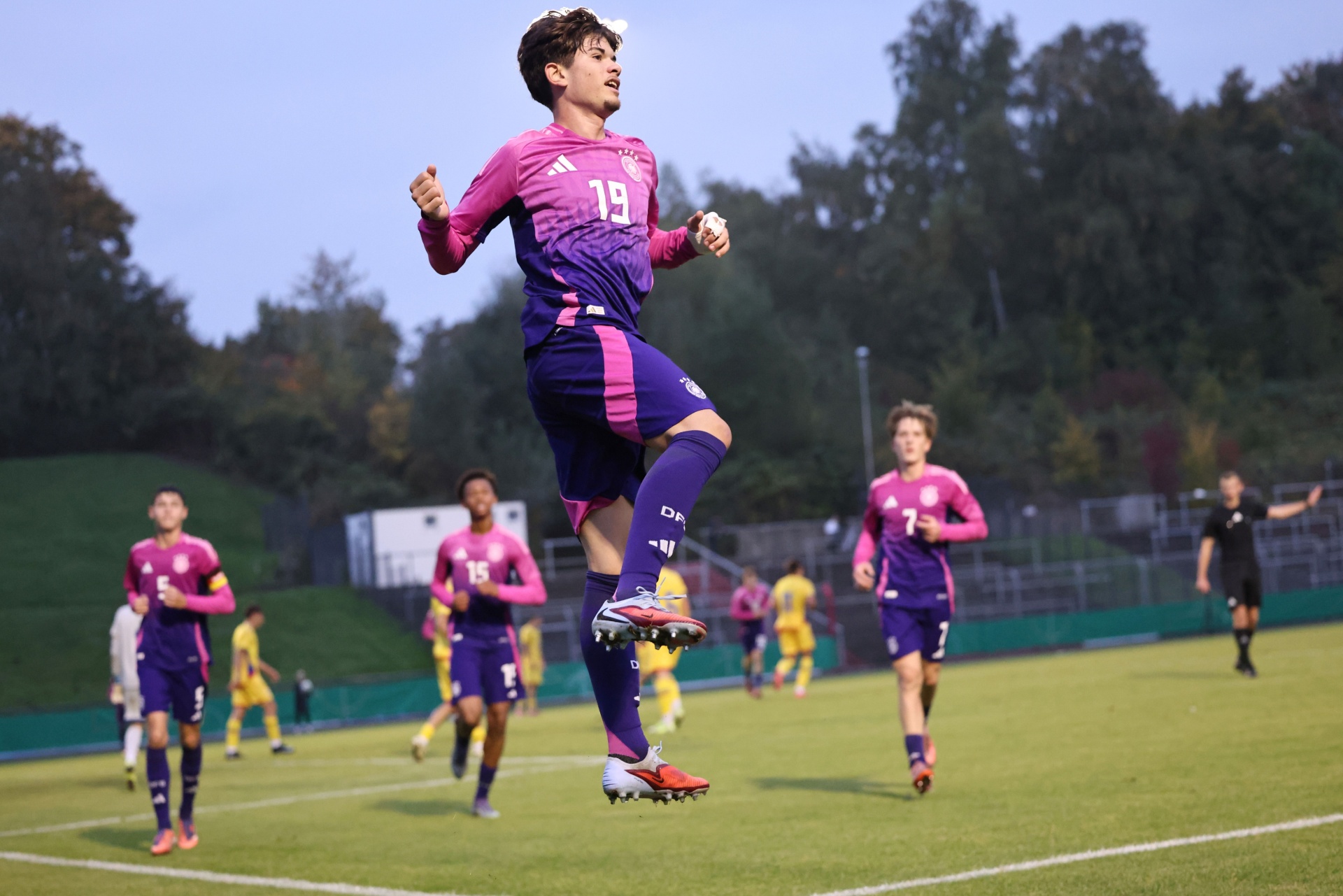 [{"clang_id":1,"value":"Fußballspieler springt jubelnd auf dem Spielfeld. Er trägt violettes Trikot mit Nummer 19 und violette Shorts. Im Hintergrund weitere Spieler und Bäume."}]