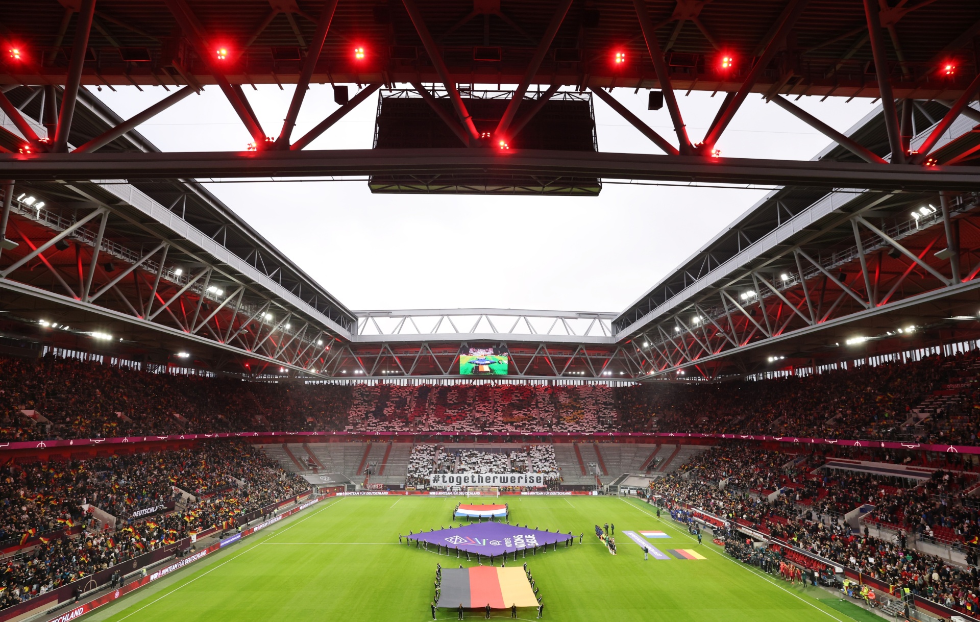 [{"clang_id":1,"value":"Fußballstadion bei Nacht: Voll besetzte Tribünen, Rasenplatz mit Spielern und Fahnen, rotes Dach mit Lichtinstallationen."}]