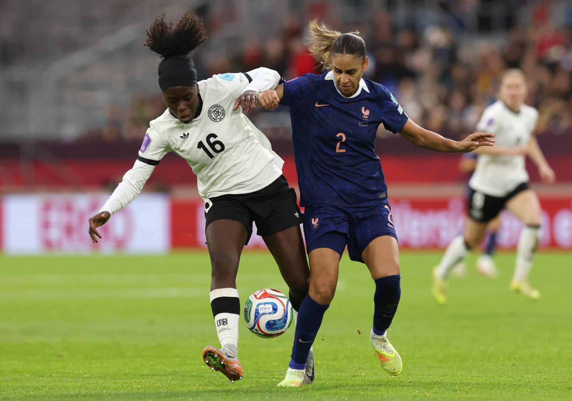 [{"clang_id":1,"value":"Zwei Fußballspielerinnen kämpfen um den Ball. Links trägt eine Spielerin den weißen Trikot mit der Nummer 16, rechts ein blaues Trikot mit der Nummer 2. Im Hintergrund weitere Spielerinnen und Zuschauer."}]