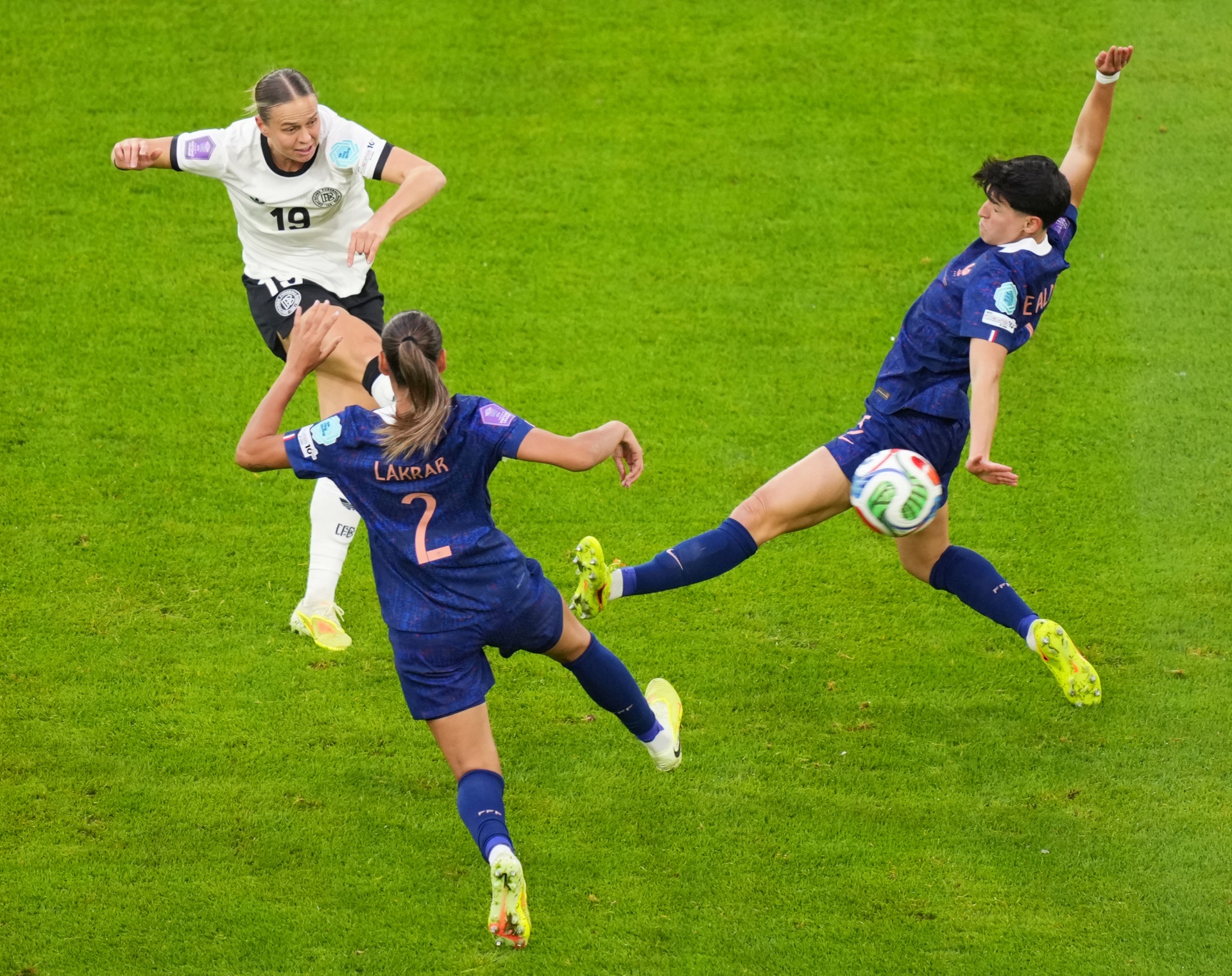 [{"clang_id":1,"value":"Drei Fußballspielerinnen auf grünem Rasen. Eine in weiß-schwarz (Nr. 19), zwei in blau (Nr. 2 und andere) – eine mit dem Fuß zum Ball, die andere mit Beinen hoch."}]