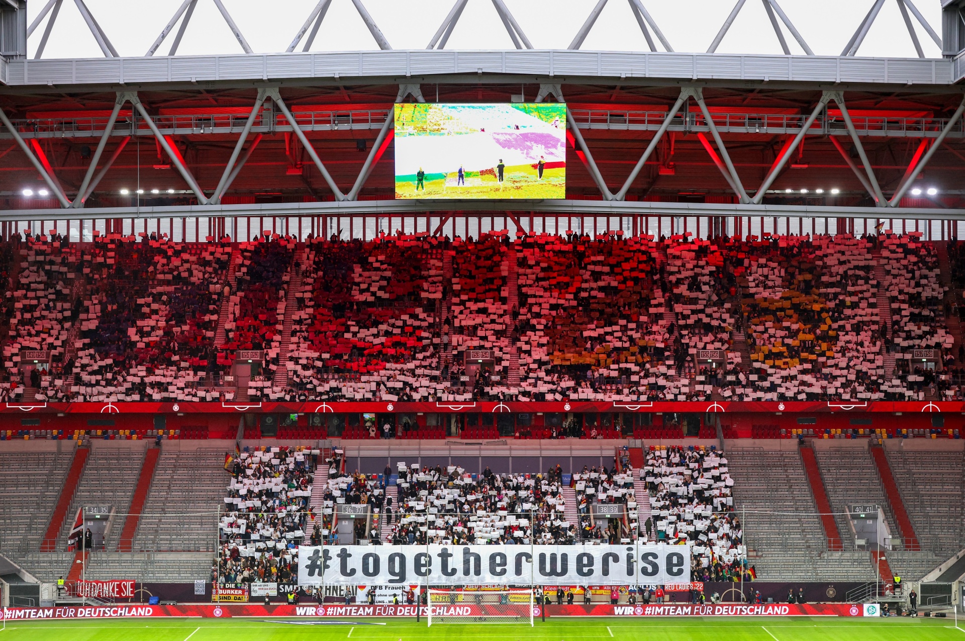 [{"clang_id":1,"value":"Fußballstadion gefüllt mit Zuschauern in rot und weiß. Eine Großbildleinwand zeigt ein Spiel. Im Vordergrund ein Banner mit der Aufschrift \"#togetherwerise\"."}]