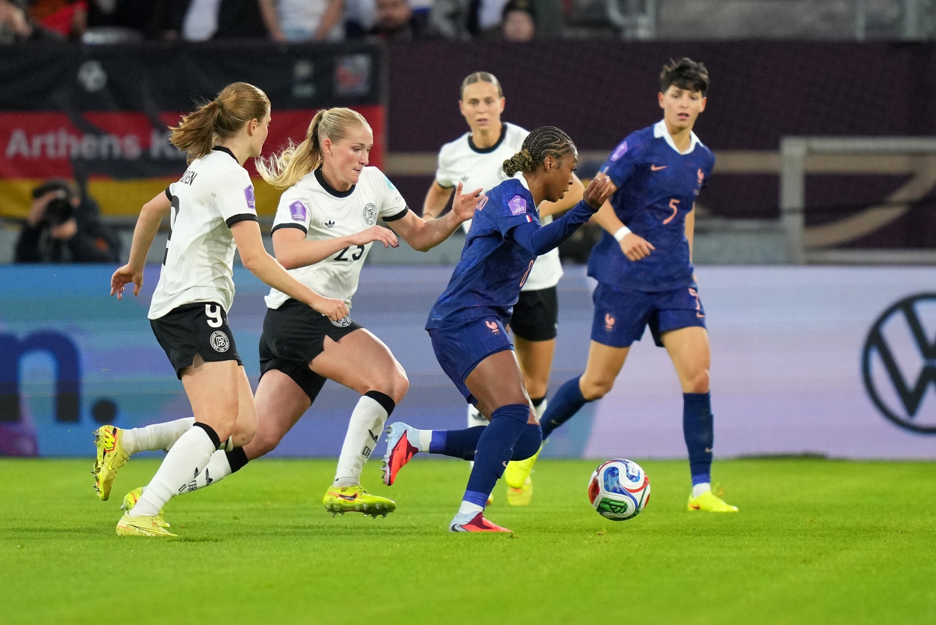 [{"clang_id":1,"value":"Fußballspiel: Spielerinnen in weißen und blauen Trikots kämpfen um den Ball auf grünem Rasen. Im Hintergrund Zuschauer und Werbebanner."}]