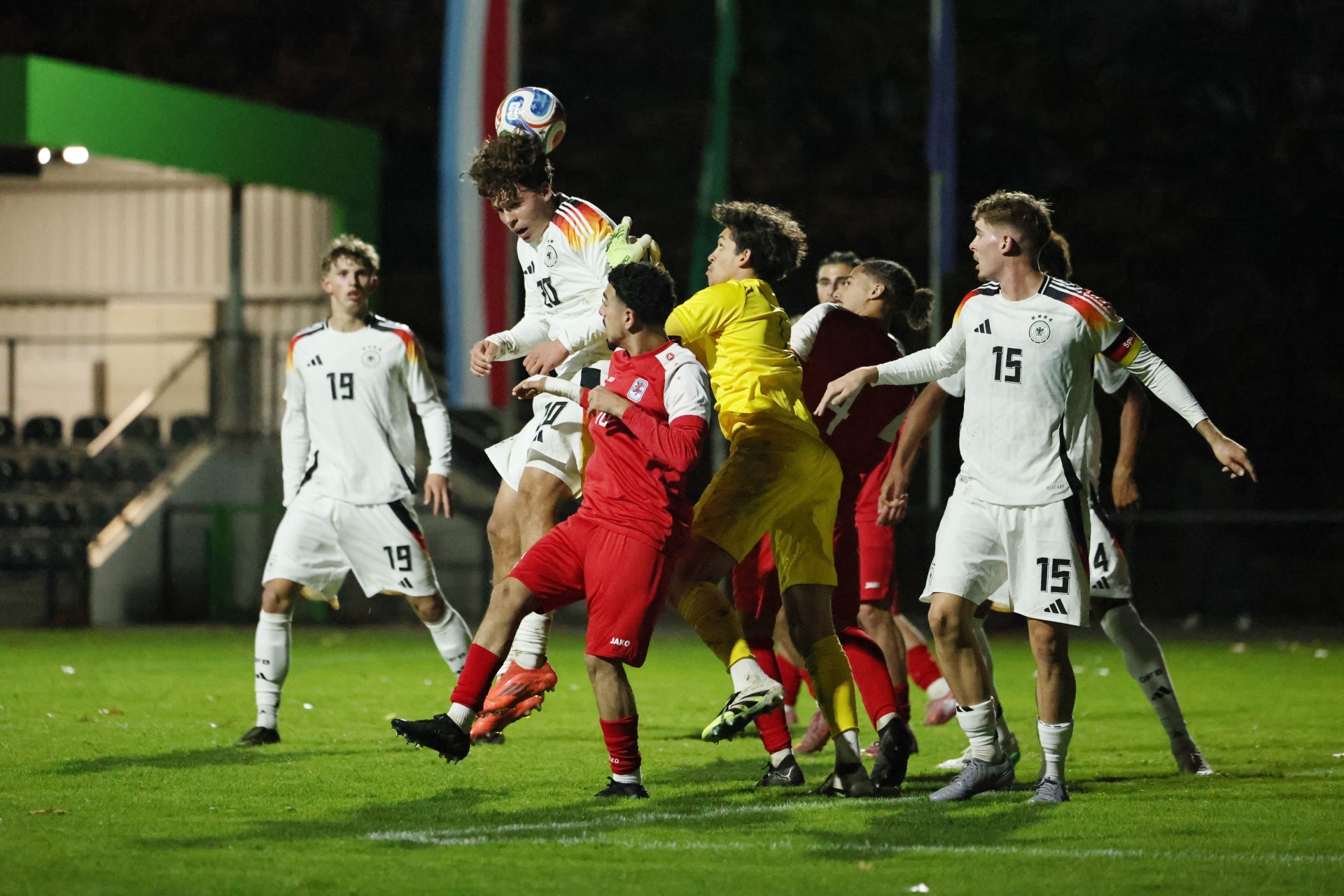 [{"clang_id":1,"value":"Fußballspiel bei Nacht. Mehrere Spieler in Rot, Gelb und Weiß kämpfen um einen Ball in der Luft. Hintergrund: Stadion."}]