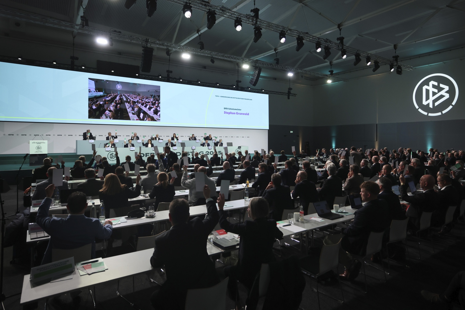 [{"clang_id":1,"value":"Großer Konferenzraum mit G20-Teilnehmern an Tischen. Im Hintergrund eine Leinwand mit Live-Bild und Namen \"Stephan Grimmel\". Beleuchtung von oben."}]