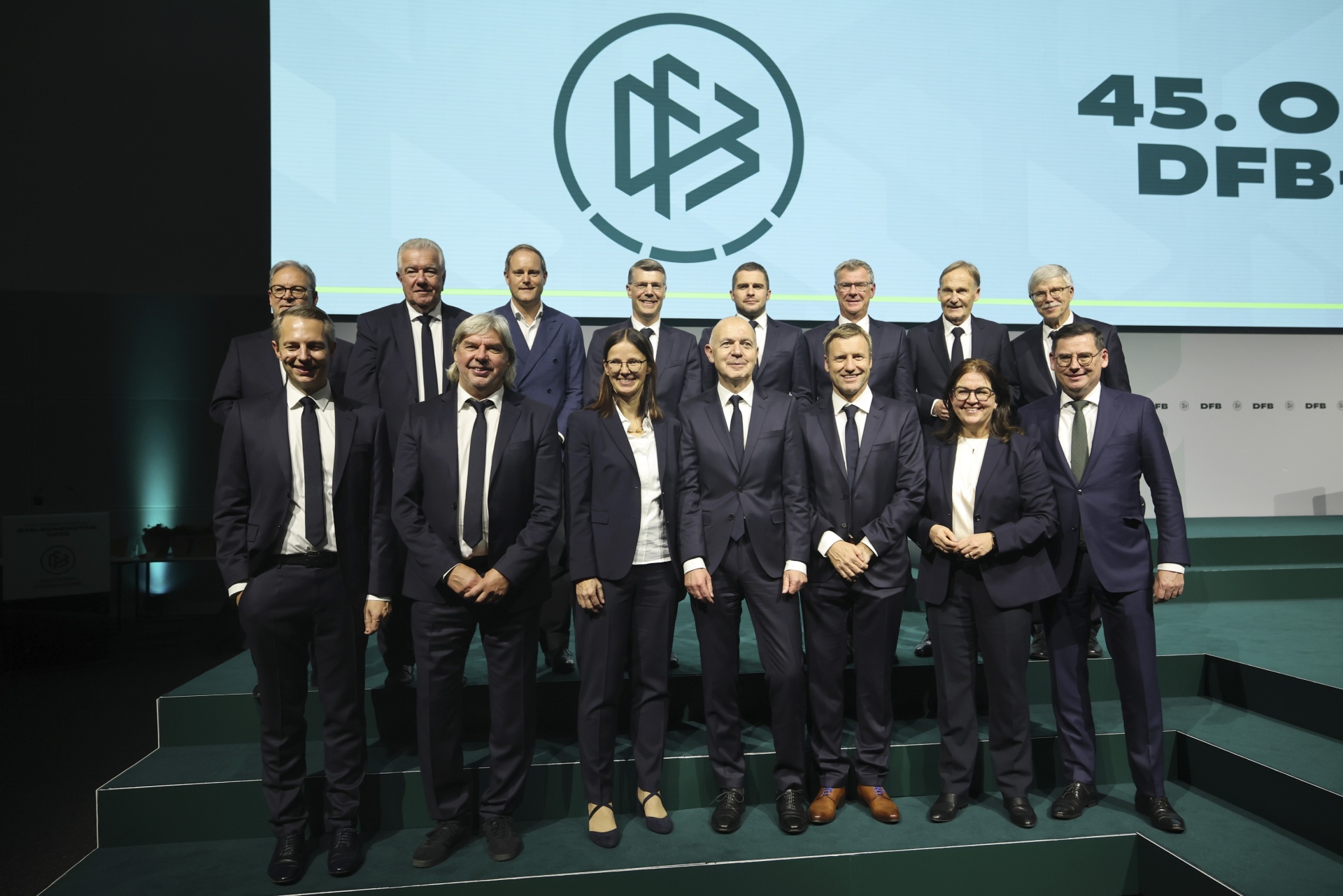 [{"clang_id":1,"value":"Gruppe von zwölf Personen in formeller Kleidung posiert vor einer grünen Wand mit dem Logo des DFB und der Aufschrift \"45.0\"."}]