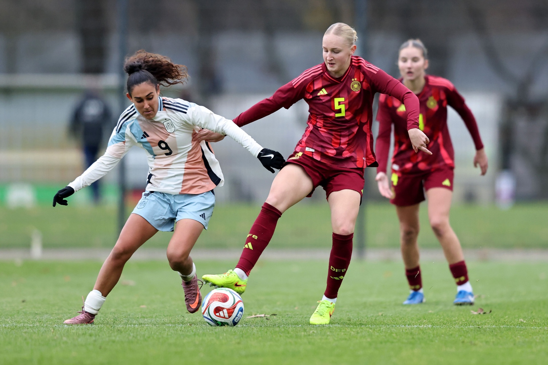 [{"clang_id":1,"value":"Zwei Fußballspielerinnen im Wettkampf. Links: Spielerin mit Nummer 9, blau-hellblaues Trikot, dribbelt den Ball. Rechts: Spielerin mit Nummer 5, bordeauxrotes Trikot, versucht den Ball zu erobern."}]
