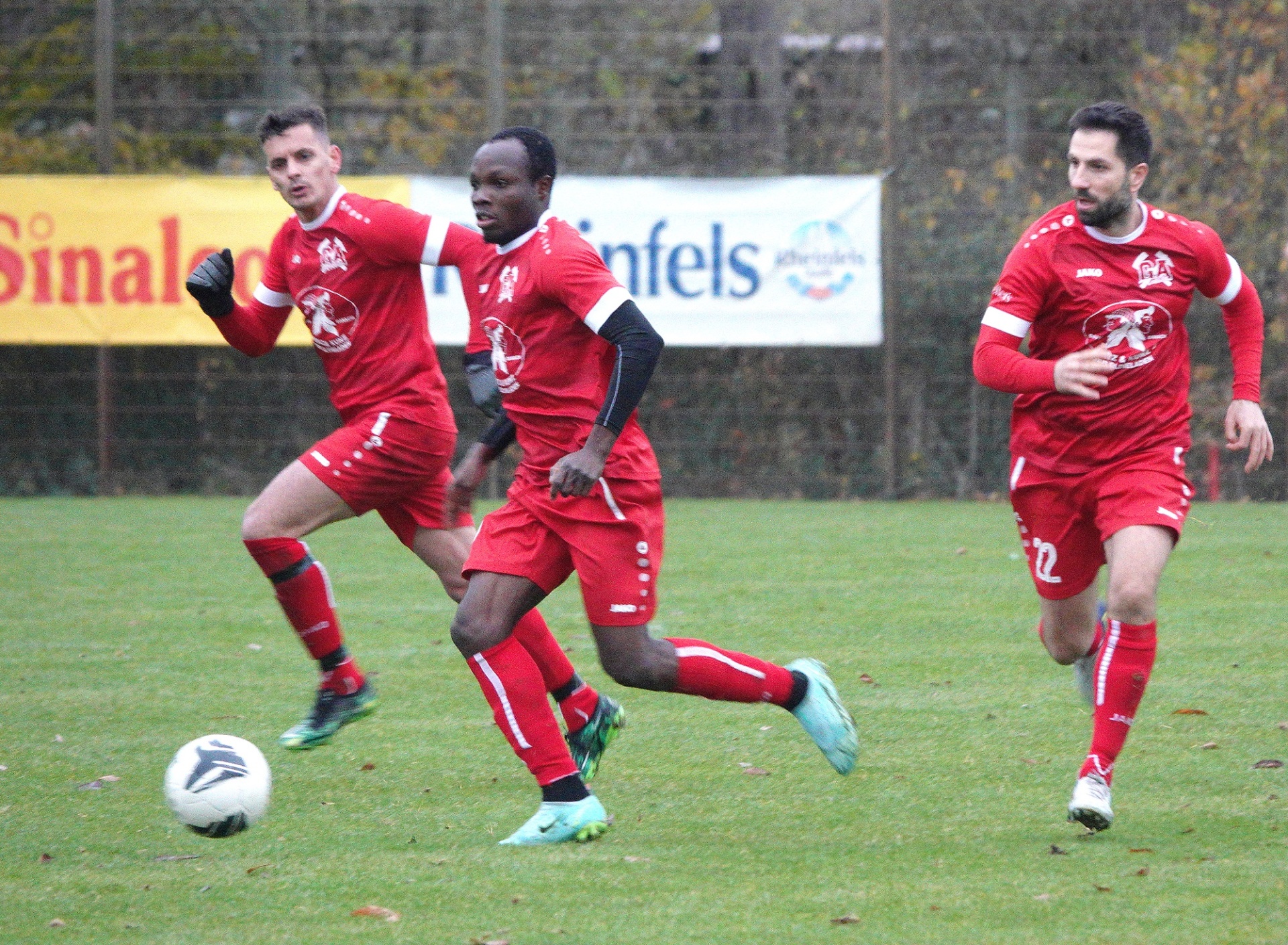 [{"clang_id":1,"value":"Drei Fußballspieler in roten Trikots laufen auf einem grünen Rasenfeld. Im Hintergrund eine metallische Zaunanlage und Werbebanner. Ein Fußball liegt am Boden."}]
