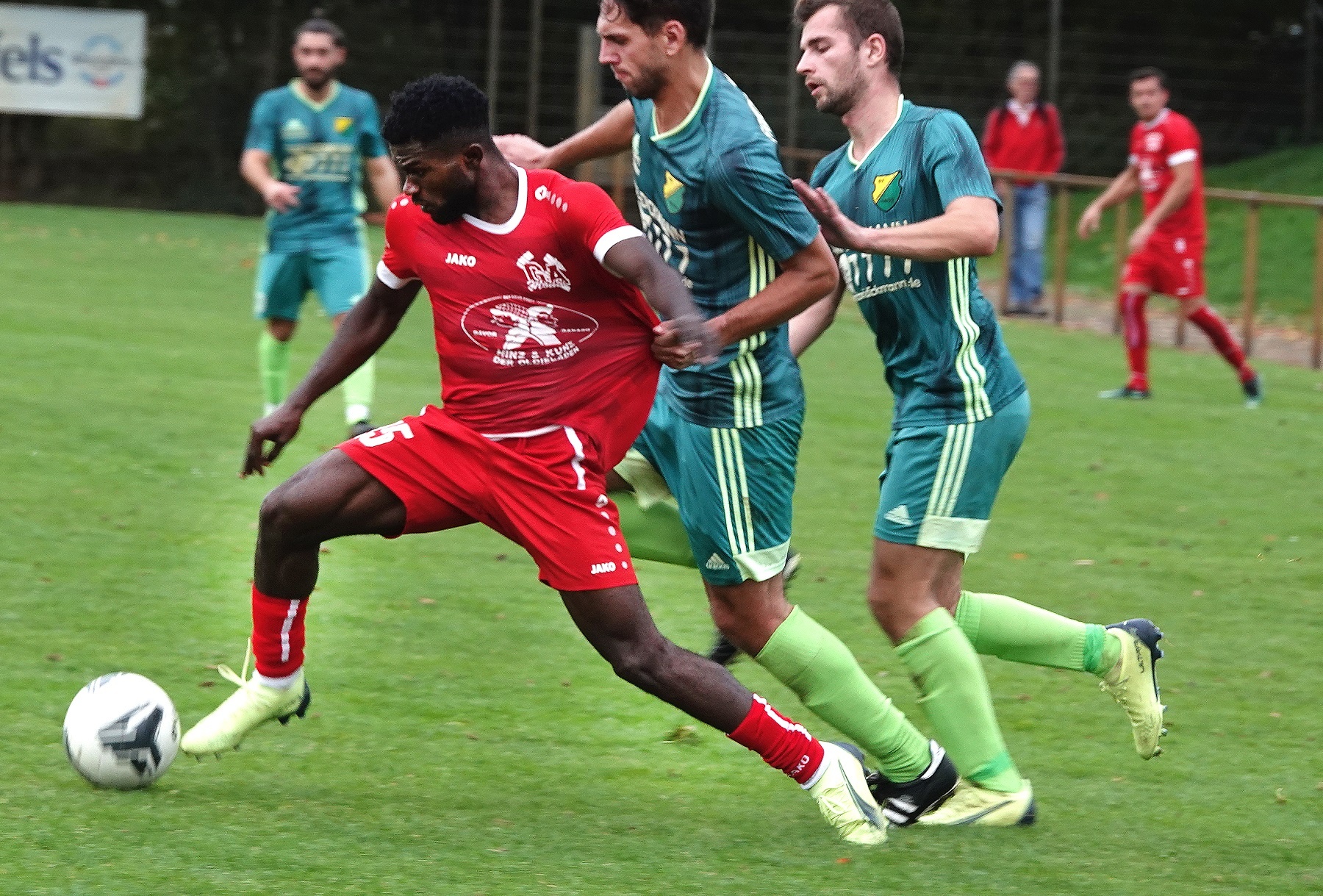 [{"clang_id":1,"value":"Fußballspiel: Spieler in roten und grünen Trikots kämpfen um den Ball auf einem Rasenplatz. Im Hintergrund Zuschauer und Bäume."}]