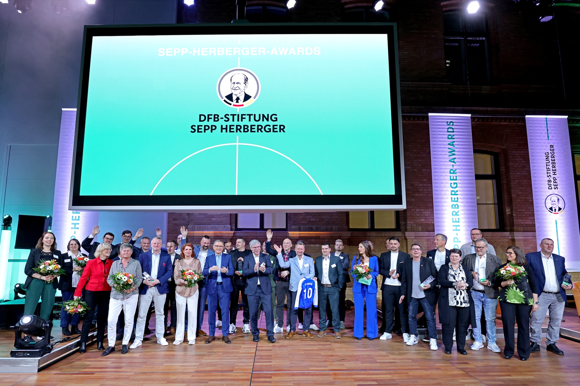 [{"clang_id":1,"value":"Gruppe von etwa 30 Personen steht auf einer Bühne vor einer großen Leinwand mit dem Text \"Sepp-Herberger-Awards\" und Logo der DFB-Stiftung Sepp Herberger."}]
