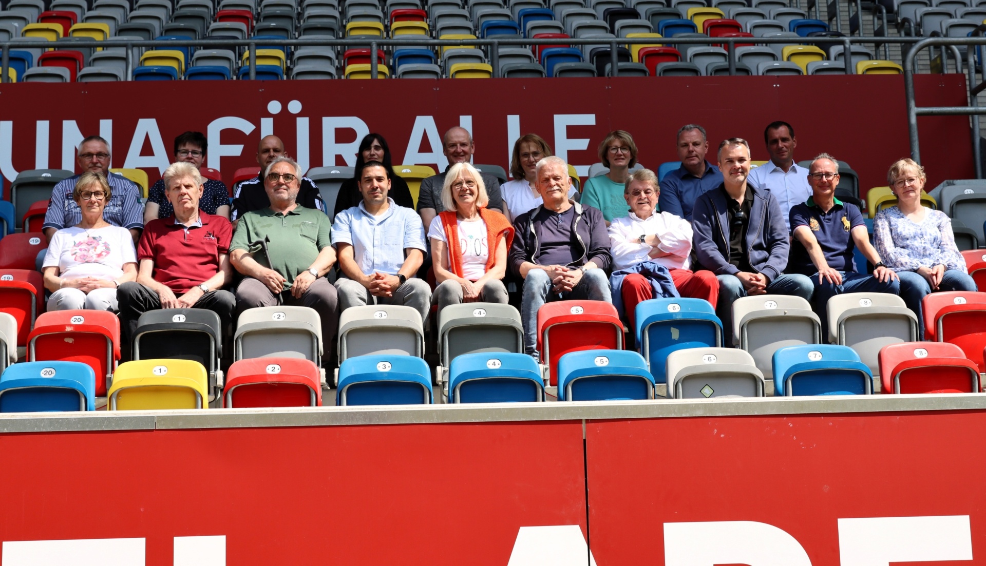 [{"clang_id":1,"value":"Zuschauer auf Stadiontribüne mit bunten Sitzen. Mehrere Personen sitzen oder stehen, dahinter weitere Zuschauerreihen. Ein Banner ist sichtbar."}]