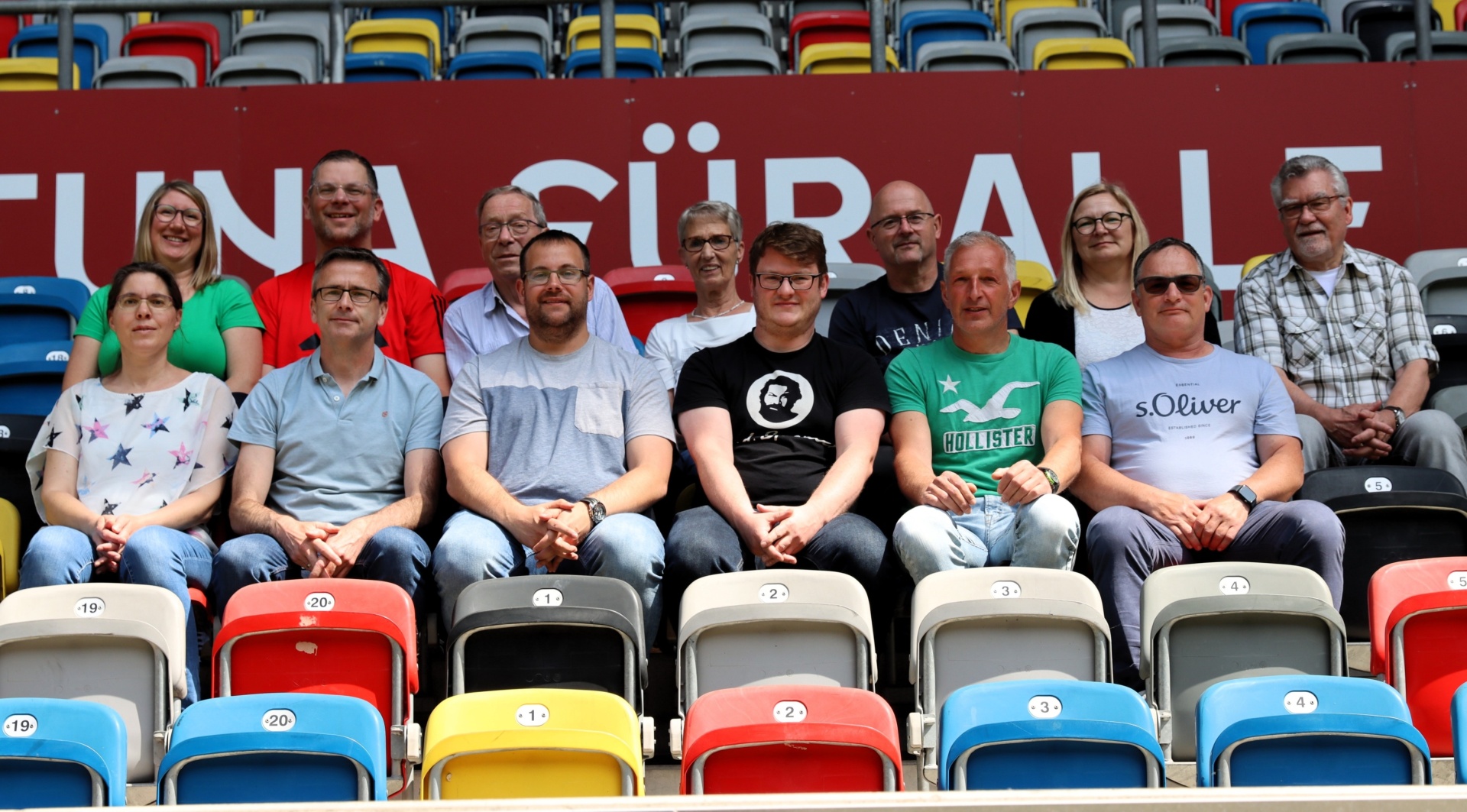 [{"clang_id":1,"value":"Zuschauerreihe in Stadion: Sieben Männer und eine Frau sitzen auf unterschiedlich farbigen Plastikstühlen. Im Hintergrund ein Banner mit Schriftzug."}]