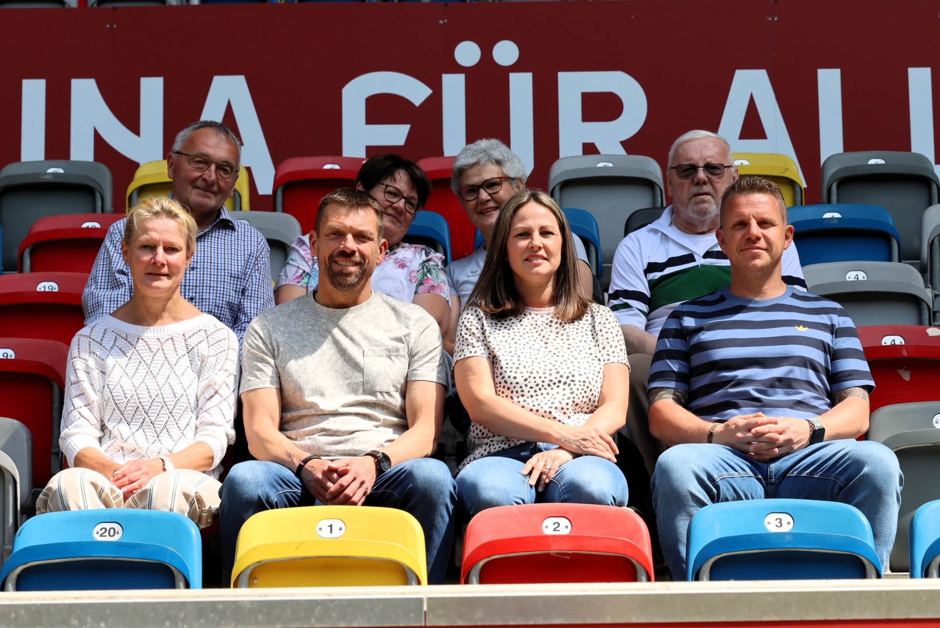 [{"clang_id":1,"value":"Zuschauerreihe in Stadion. Sechs Personen sitzen auf farbigen Sitzen, im Hintergrund ein Banner mit großem Text."}]