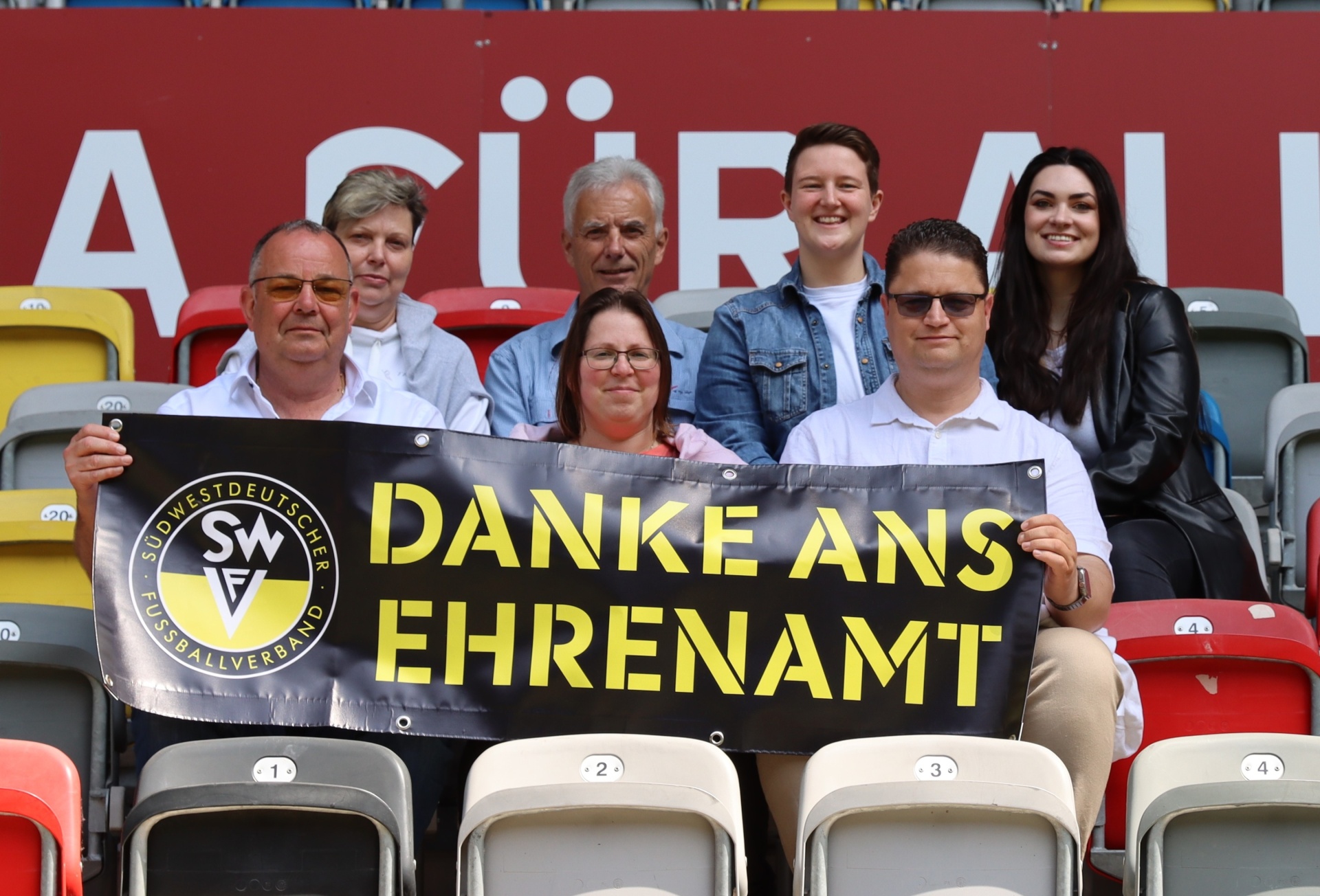 [{"clang_id":1,"value":"Gruppe von acht Personen sitzt auf Stadiontribünen. Sie halten ein Banner mit der Aufschrift \"DANKE ANS EHRENAMT\" und dem Logo des Südwestdeutschen Fußballverbands."}]
