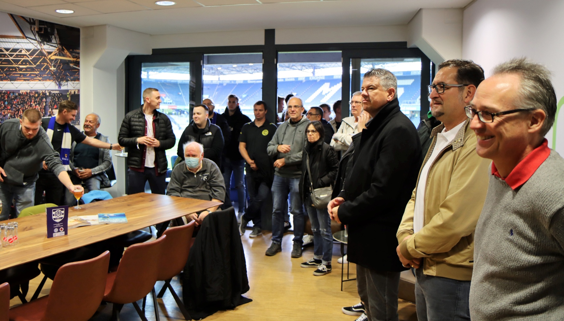 [{"clang_id":1,"value":"Eine Gruppe von etwa 20 Personen steht und sitzt in einem Raum mit Blick auf ein Fußballstadion. Mehrere tragen dunkle Kleidung, einige haben Brillen."}]