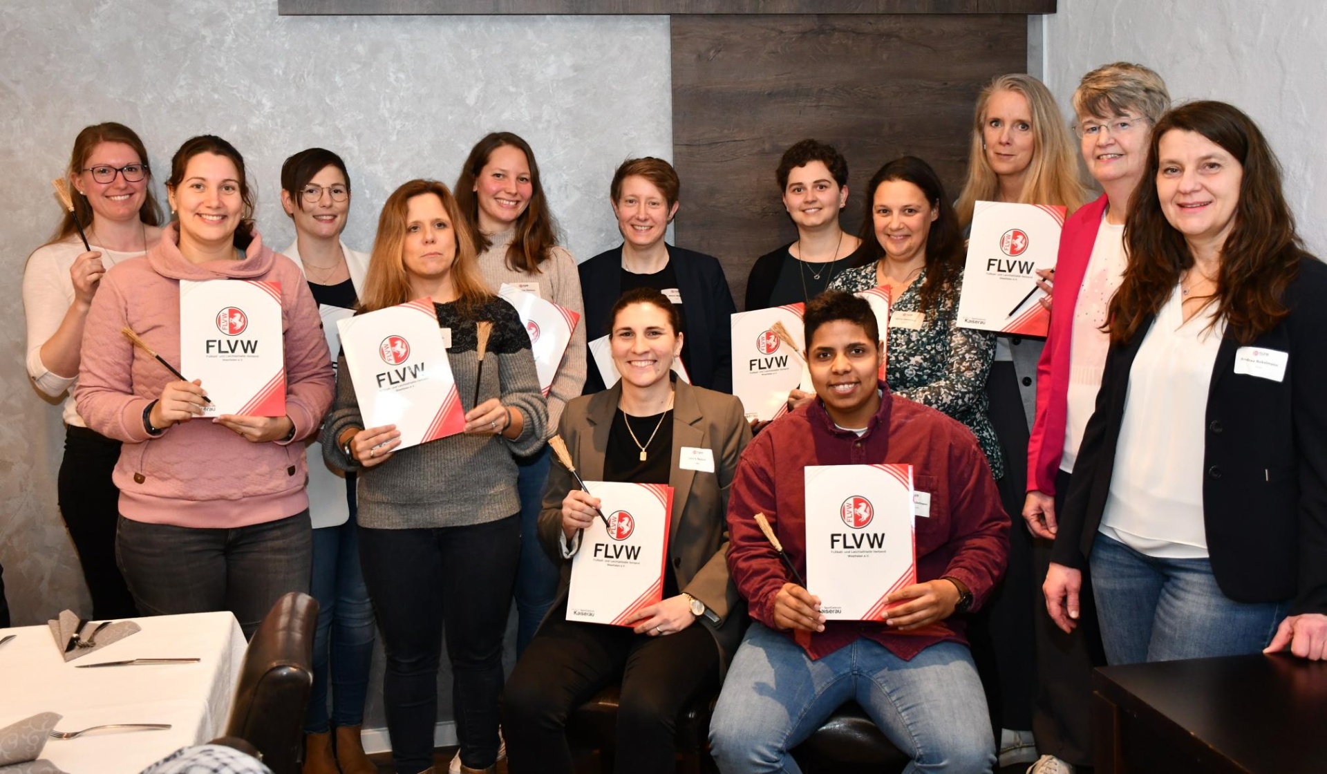 [{"clang_id":1,"value":"Gruppe von 16 Personen, überwiegend Frauen, hält Zertifikate mit dem Logo \"FLVW\" in den Händen. Sie stehen vor einer hellen Wand."}]
