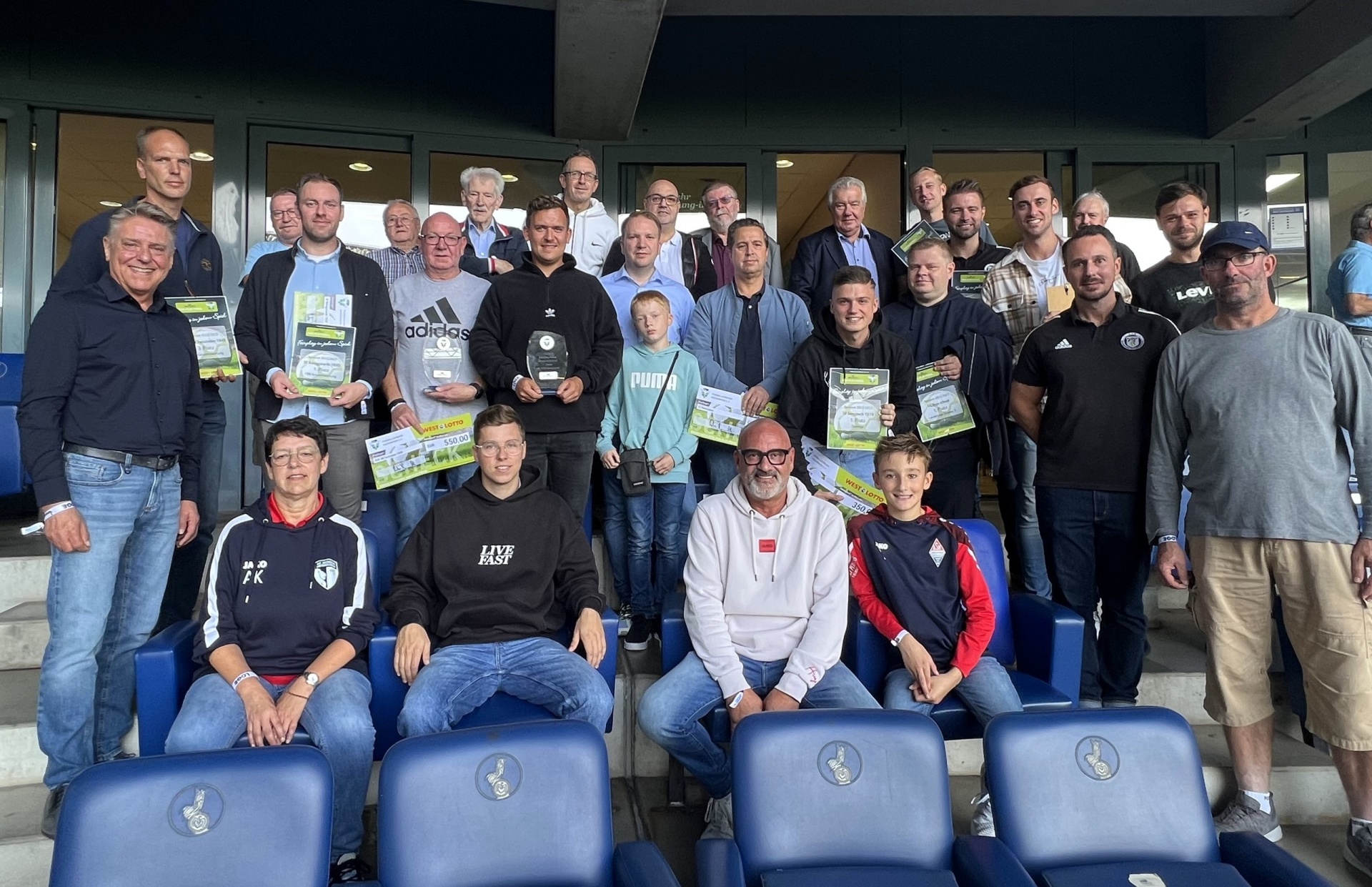 [{"clang_id":1,"value":"Eine Gruppe von etwa 30 Männern und wenigen Frauen sitzt oder steht auf blauen Stadion-Sesseln. Viele halten Flyer oder Programmhefte. Im Hintergrund eine Glasfront."}]