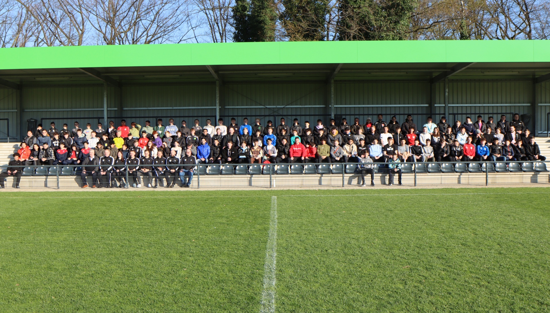 [{"clang_id":1,"value":"Mannschaftsfoto auf Stadiontribüne. Eine große Gruppe von Personen sitzt auf einer Bank vor einem Stadiondach, vor ihnen ein Fußballfeld."}]