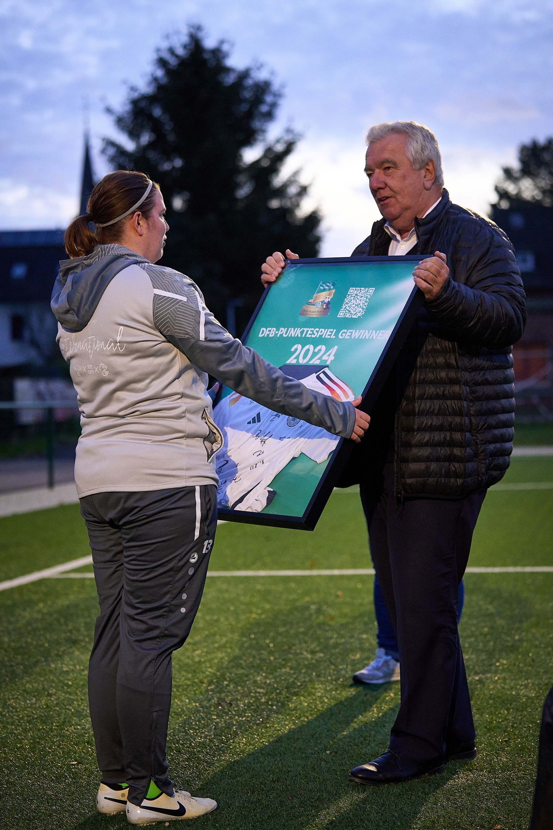 [{"clang_id":1,"value":"Eine Frau in Sportkleidung nimmt von einem Mann in Jacke eine gerahmte Plakette mit Aufschrift \"DFB-Punktespiel Gewinn 2024\" entgegen. Hintergrund: Fußballfeld und Bäume."}]