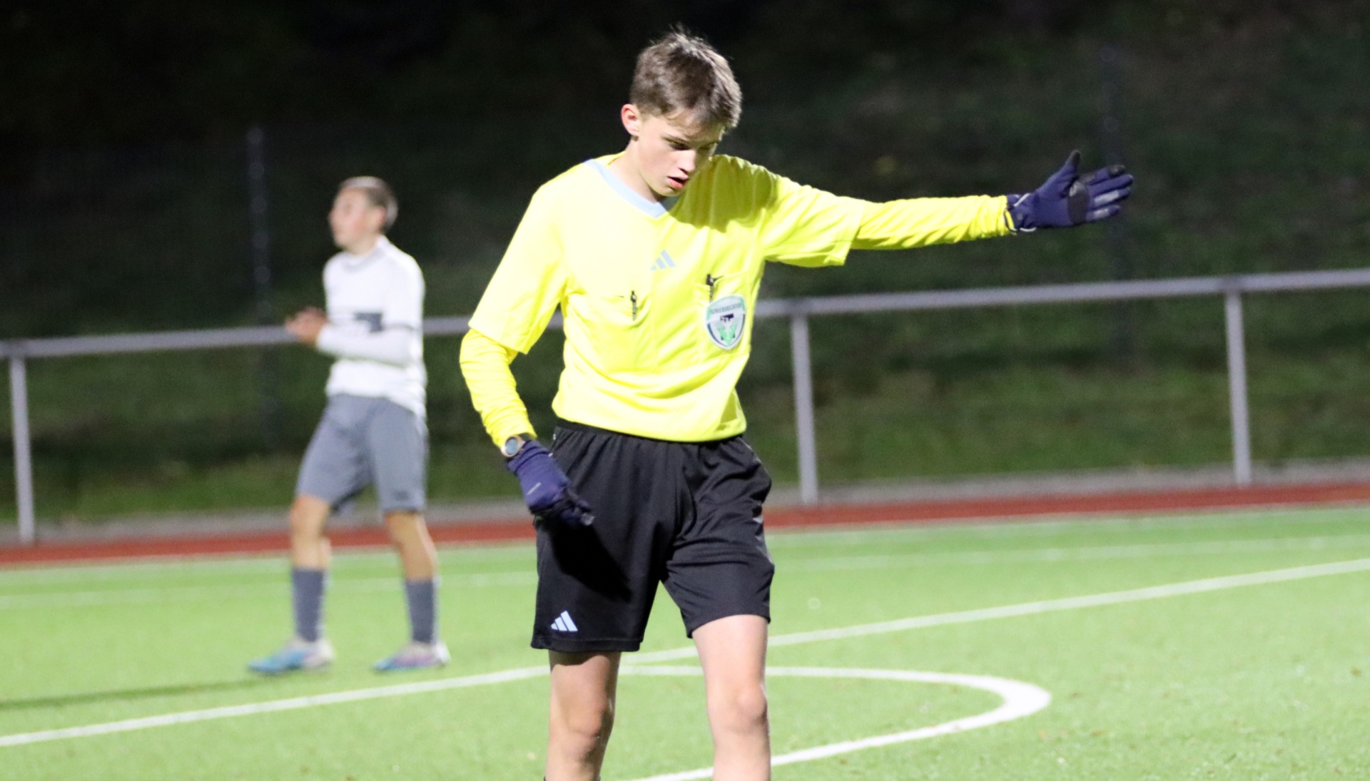 [{"clang_id":1,"value":"Fußballtorwart in gelbem Trikot und schwarzen Shorts auf grünem Rasen, Arm ausgestreckt. Im Hintergrund ein weiterer Spieler und eine Absperrung."}]