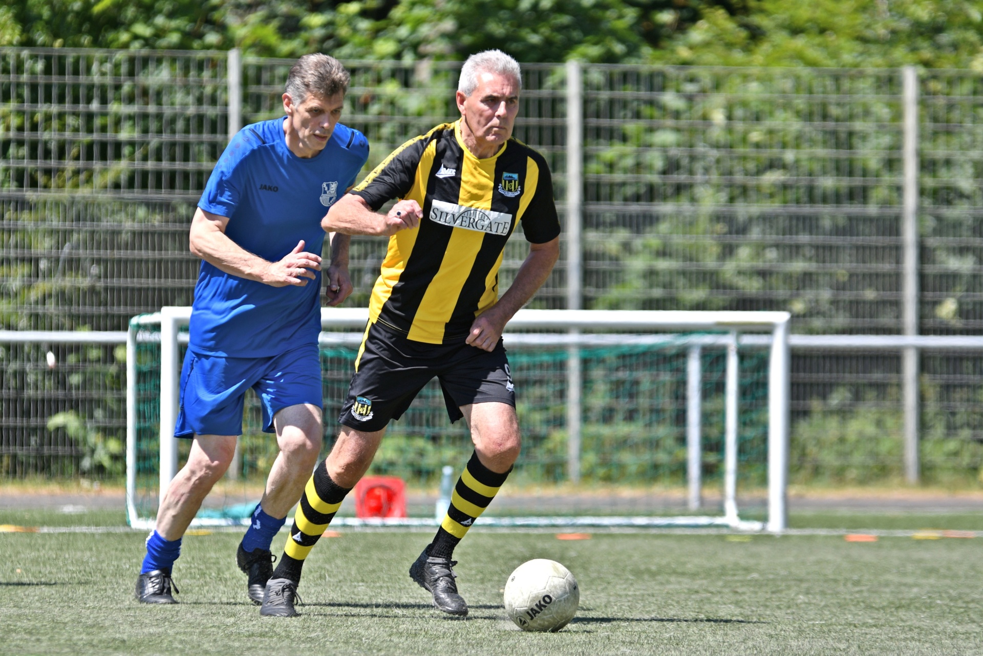 [{"clang_id":1,"value":"Zwei Männer spielen Fußball auf einem Kunstrasenplatz. Der eine trägt blaues Trikot und Shorts, der andere schwarz-gelbes Trikot mit Sponsorlogo und schwarzen Shorts."}]