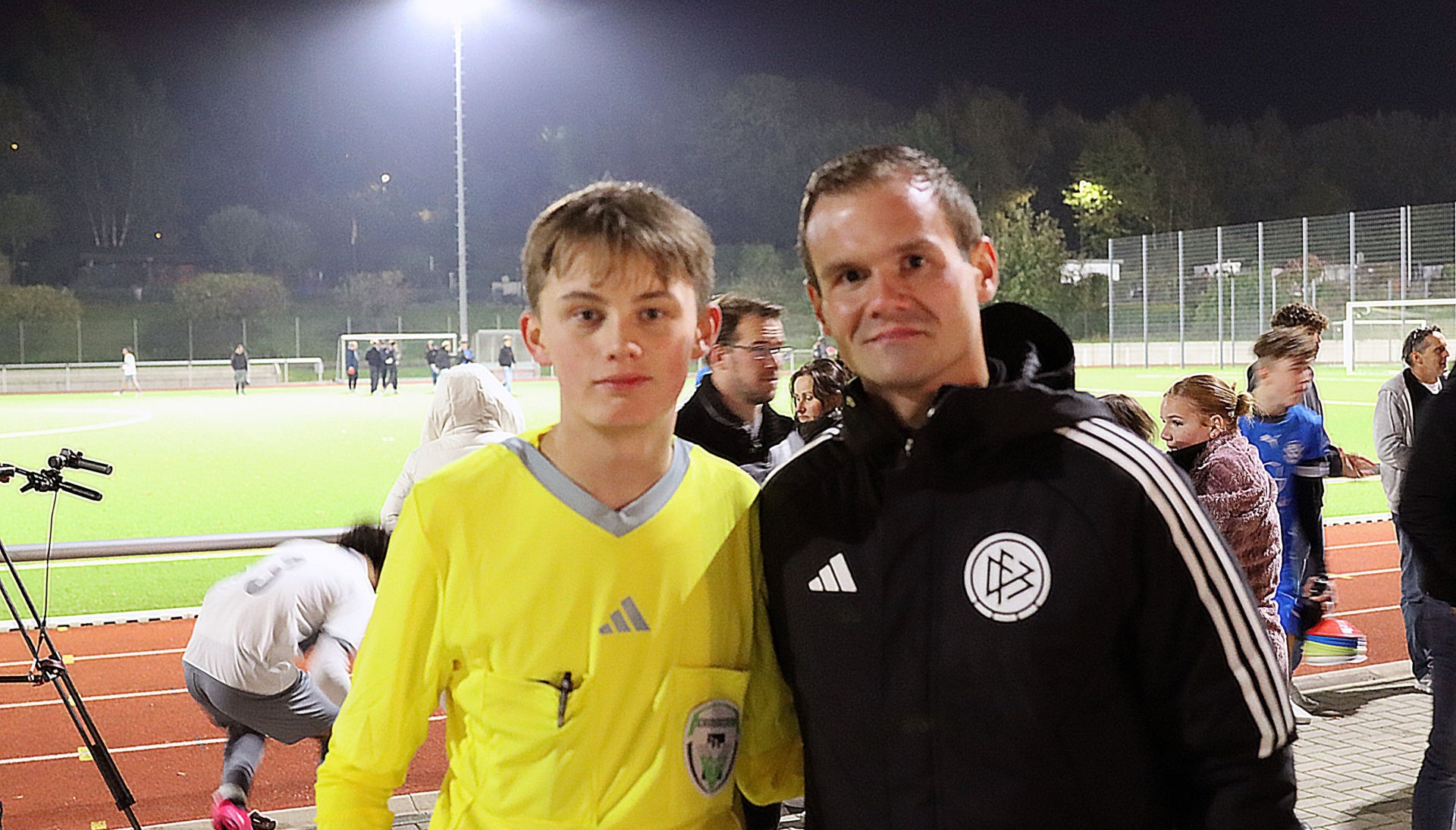 [{"clang_id":1,"value":"Zwei Männer posieren vor einer Fußballfeldbeleuchtung. Links ein Jugendlicher im gelben Trikot, rechts ein Mann in schwarzer Sportjacke mit Logo. Im Hintergrund verschwommen Zuschauer."}]