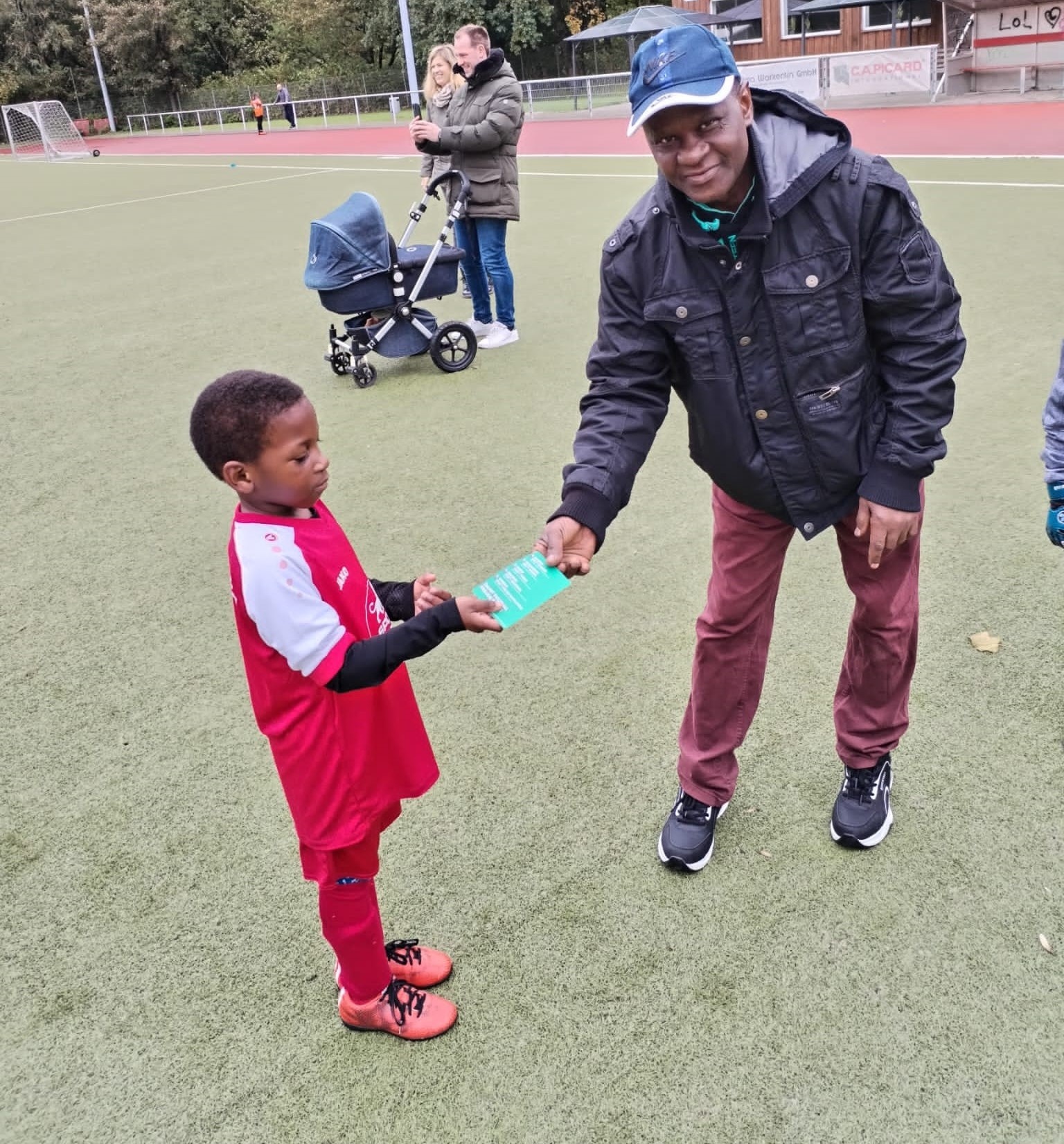 [{"clang_id":1,"value":"Junge im Fußballtrikot gibt einem Mann die Hand. Hintergrund: Sportplatz mit Zuschauern und Gebäuden."}]