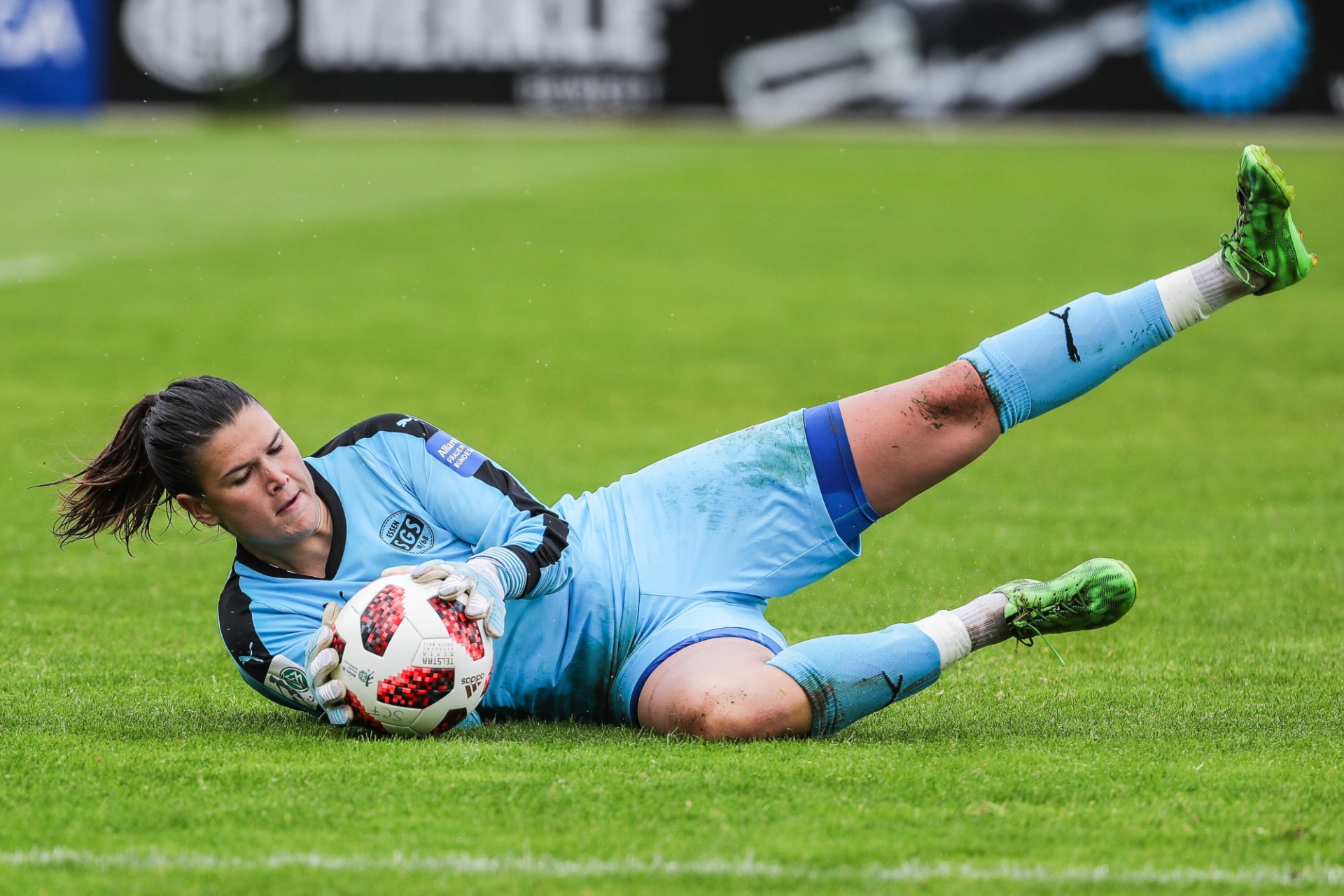 [{"clang_id":1,"value":"Fußballtorhüterin in blauer Ausrüstung und grünen Schuhen während einer Parade auf grünem Rasen. Sie hält einen Fußball fest."}]