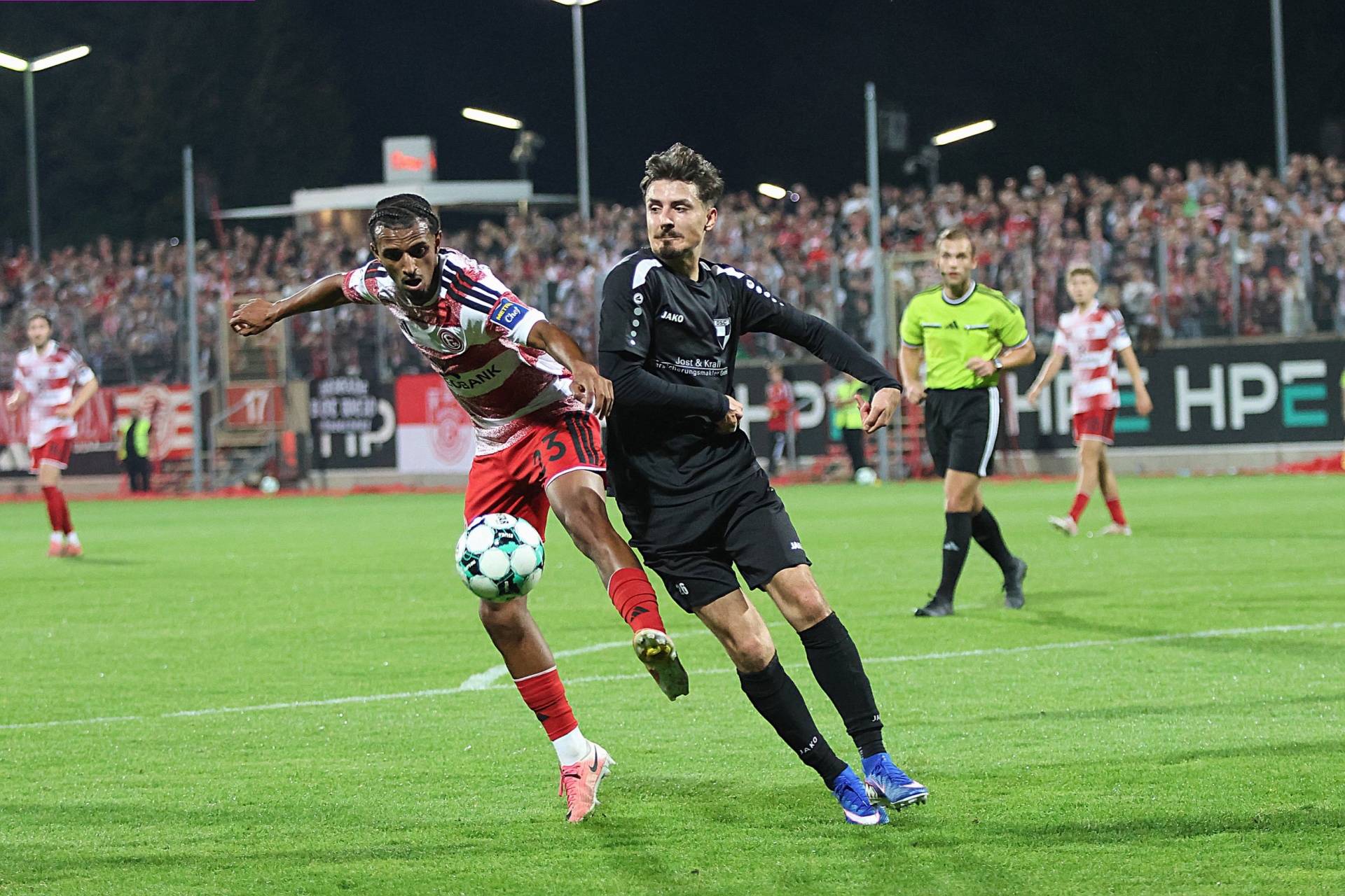 Zwei Fußballspieler im Spiel: Ein Spieler mit rotem Trikot tritt den Ball, ein Spieler mit schwarzem Trikot verteidigt. Im Hintergrund Zuschauer und Flutlicht.