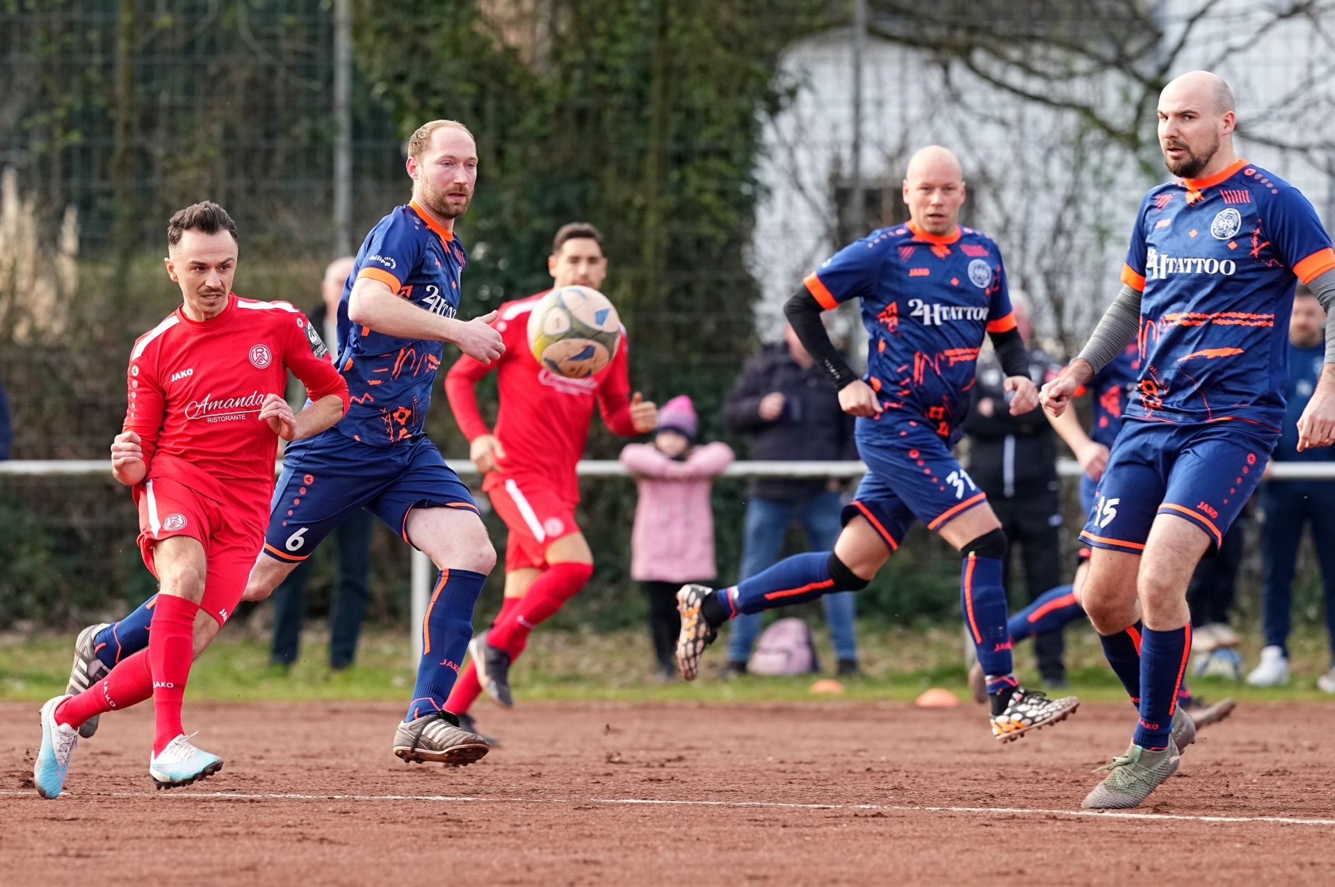 [{"clang_id":1,"value":"Fußballspiel auf Rasenplatz. Mehrere Spieler in roten und blau-navy Trikots im Wettkampf. Zuschauer im Hintergrund."}]