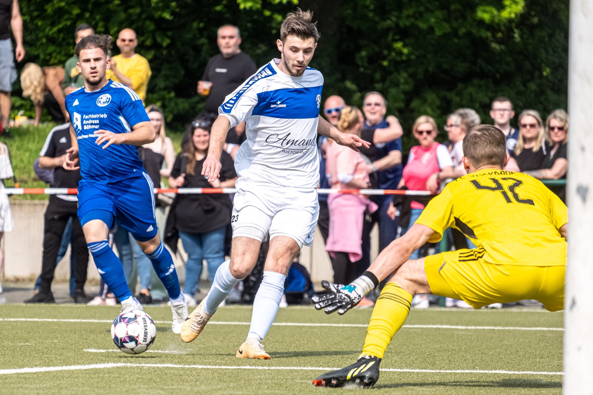 [{"clang_id":1,"value":"Fußballspiel: Spieler in blauem Trikot schießt auf Tor, gelber Torwart in Aktion. Im Hintergrund Zuschauer. Rasenplatz."}]