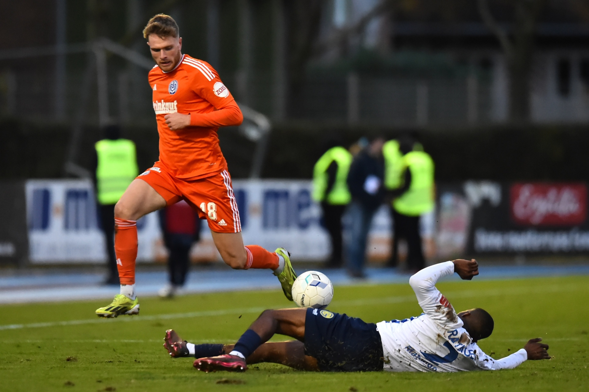 [{"clang_id":1,"value":"Fußballspieler in orange und blau-weiß rutschen auf Rasen. Der orange gekleidete Spieler tritt den Ball. Im Hintergrund Zuschauer und Stadion."}]