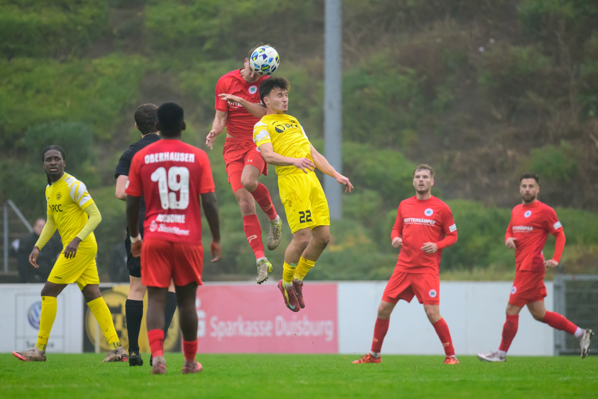 [{"clang_id":1,"value":"Fußballspiel: Zwei Spieler springen um den Ball. Im Hintergrund weitere Spieler und eine Werbebande mit dem Schriftzug \"Sparkasse Duisburg\". Rasenplatz."}]