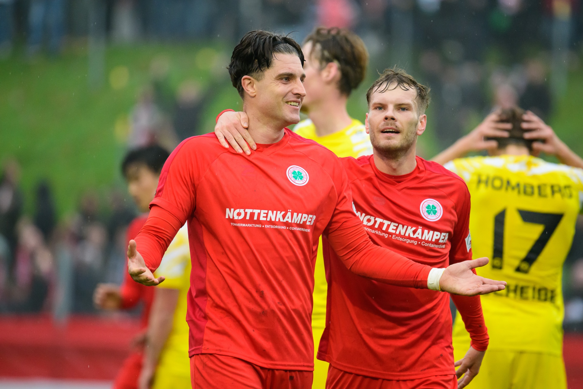[{"clang_id":1,"value":"Zwei Fußballspieler in roten Trikots mit grünen Akzenten stehen eng nebeneinander und lächeln in die Kamera. Im Hintergrund unscharfe Spieler in gelben Trikots."}]