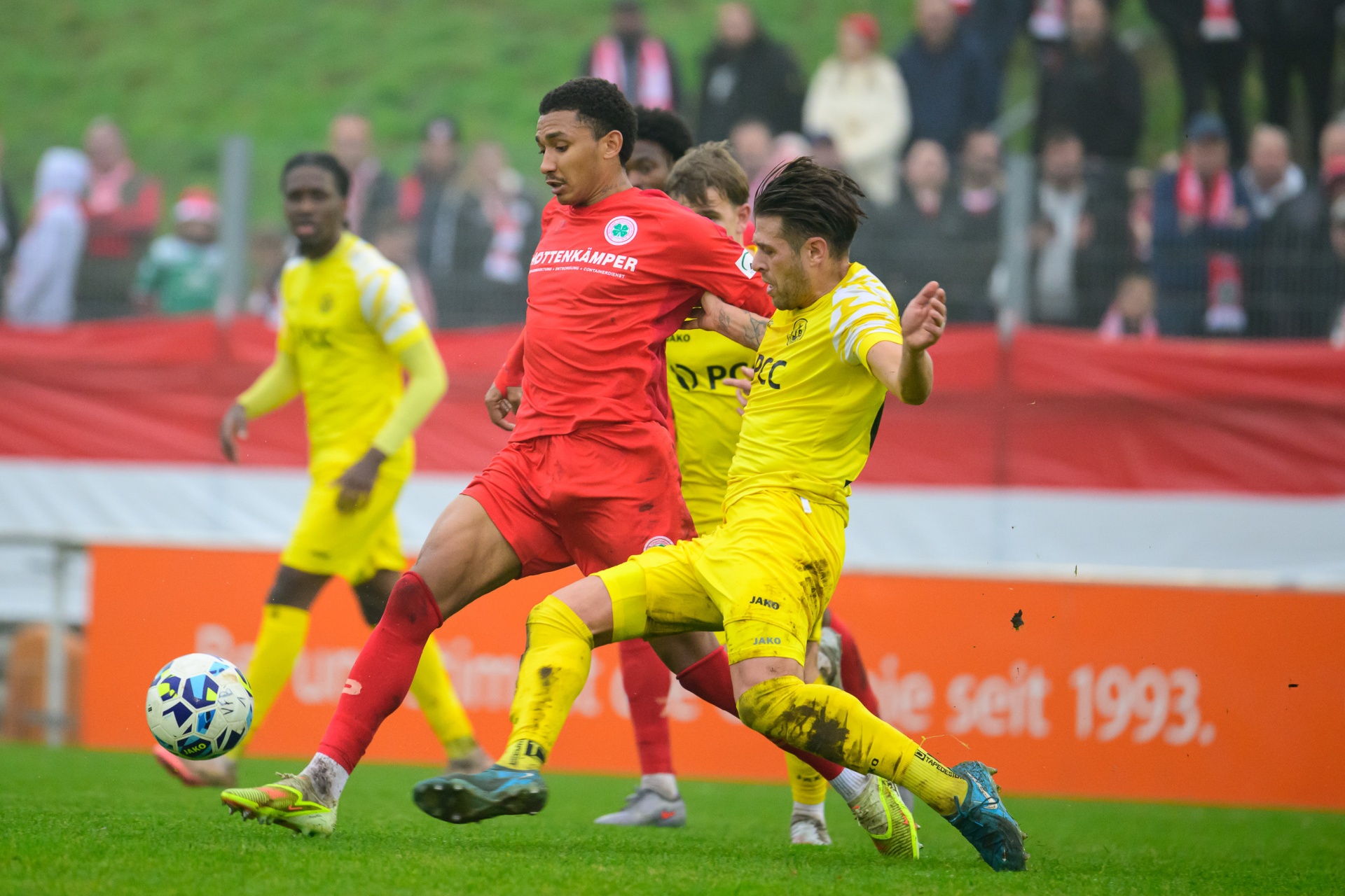 [{"clang_id":1,"value":"Zwei Fußballspieler kämpfen um den Ball auf grünem Rasen. Links ein Spieler in rotem Trikot, rechts in gelb. Im Hintergrund weitere Spieler und Zuschauer."}]