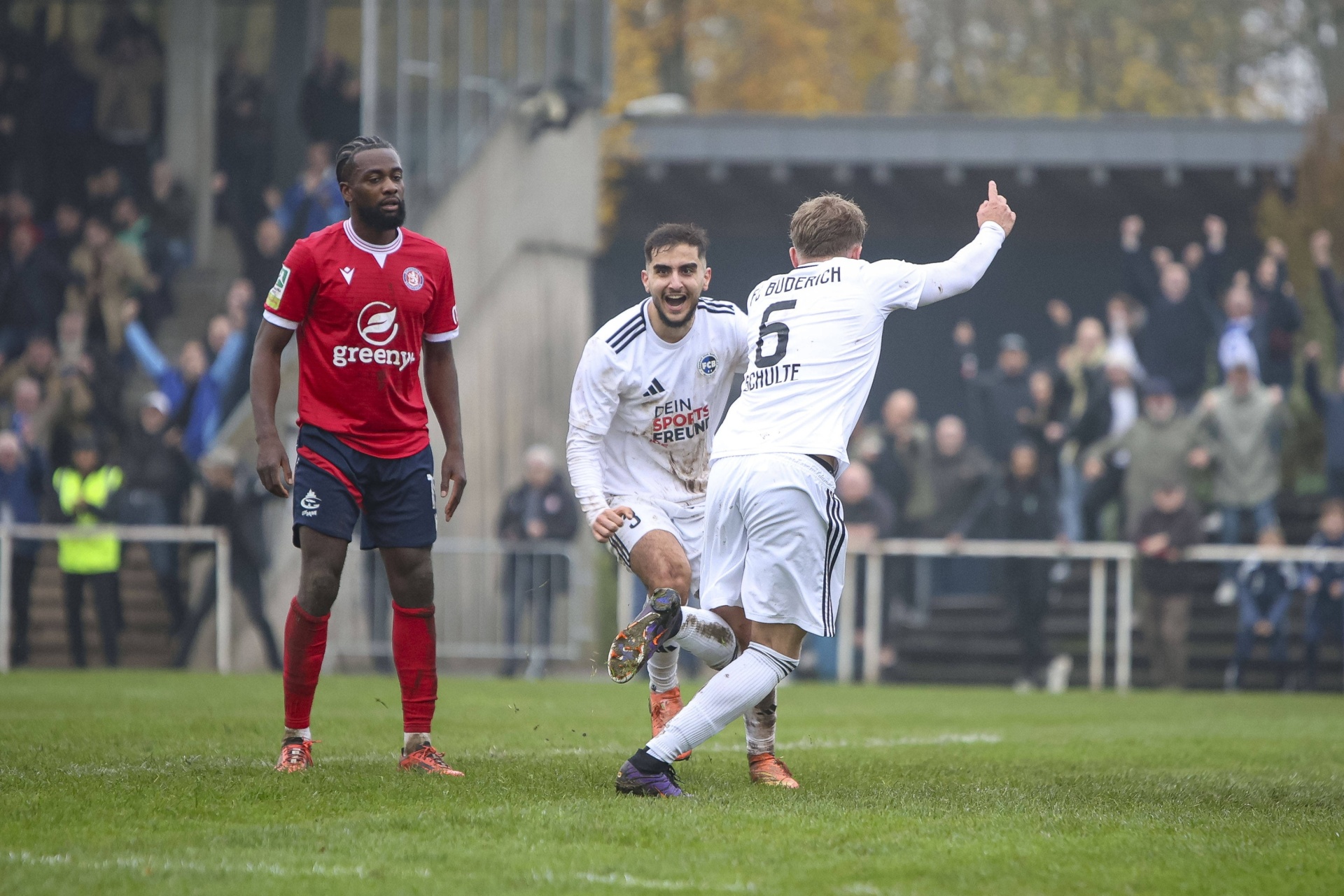 [{"clang_id":1,"value":"Drei Fußballspieler auf einem Rasenplatz. Links ein Spieler mit rotem Trikot, in der Mitte und rechts zwei Spieler mit weißen Trikots, einer jubelt. Hintergrund: Zuschauer und ein Zaun."}]