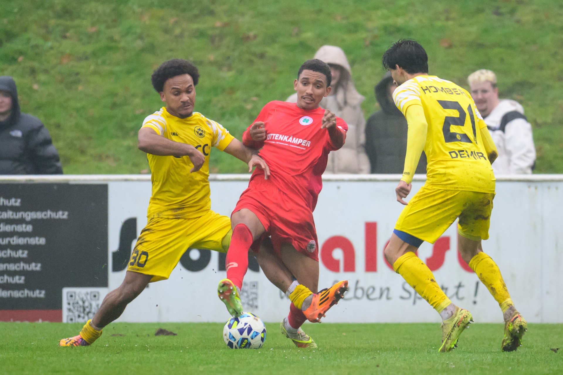 [{"clang_id":1,"value":"Drei Fußballspieler auf grünem Rasen. Links und rechts Spieler in gelben Trikots, mittig ein Spieler in rotem Trikot mit Ball. Im Hintergrund Werbebanner."}]