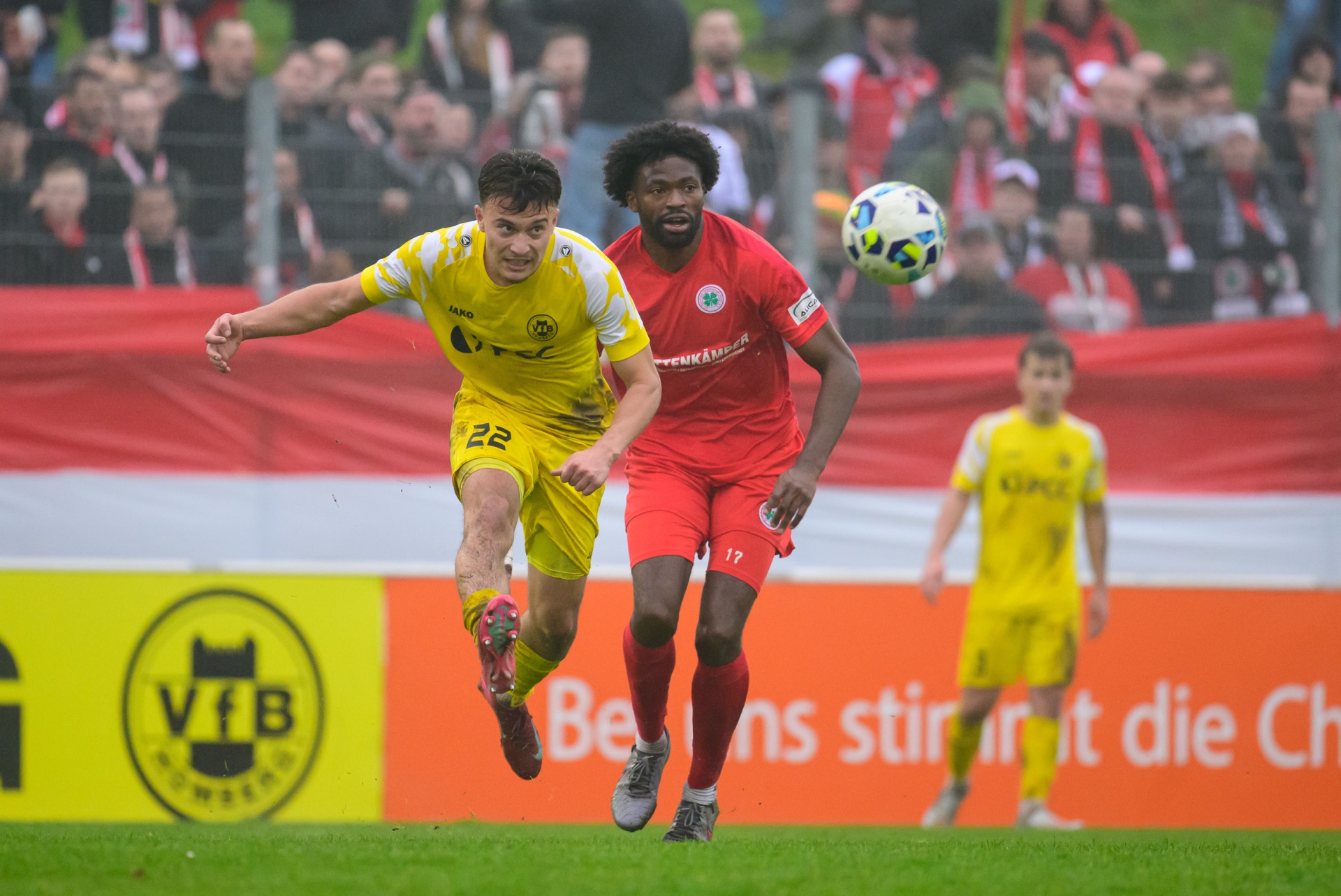 [{"clang_id":1,"value":"Fußballspiel: Zwei Spieler, einer in Gelb (Nr. 22) und einer in Rot (Nr. 17), kämpfen um den Ball. Im Hintergrund Zuschauer und ein Stadionbanner."}]