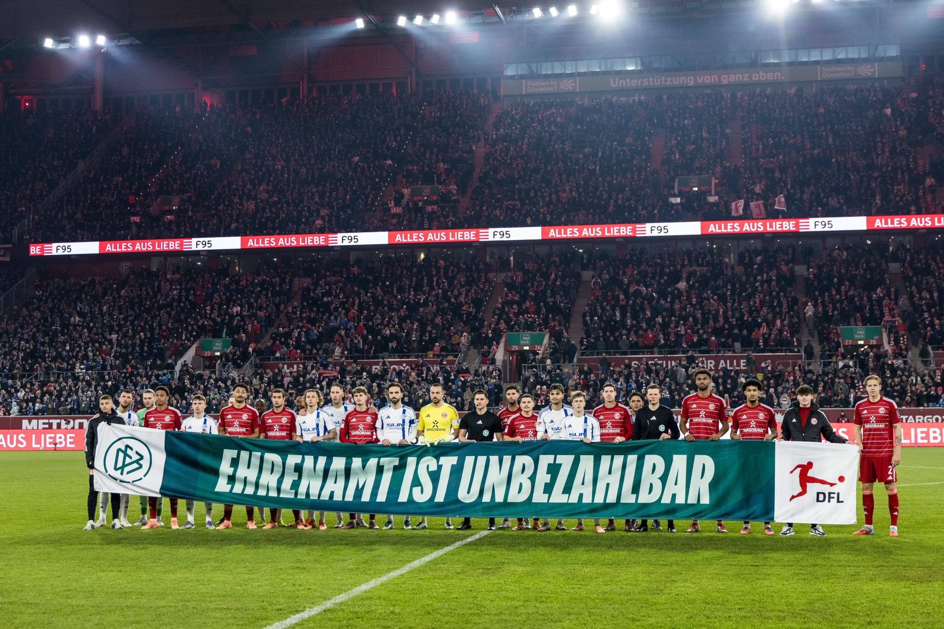 [{"clang_id":1,"value":"Fußballstadion gefüllt mit Zuschauern. Spieler stehen auf dem Rasen vor einem Banner mit dem Text \"Ehrenamt ist unbezahlbar\" und DFL-Logo."}]