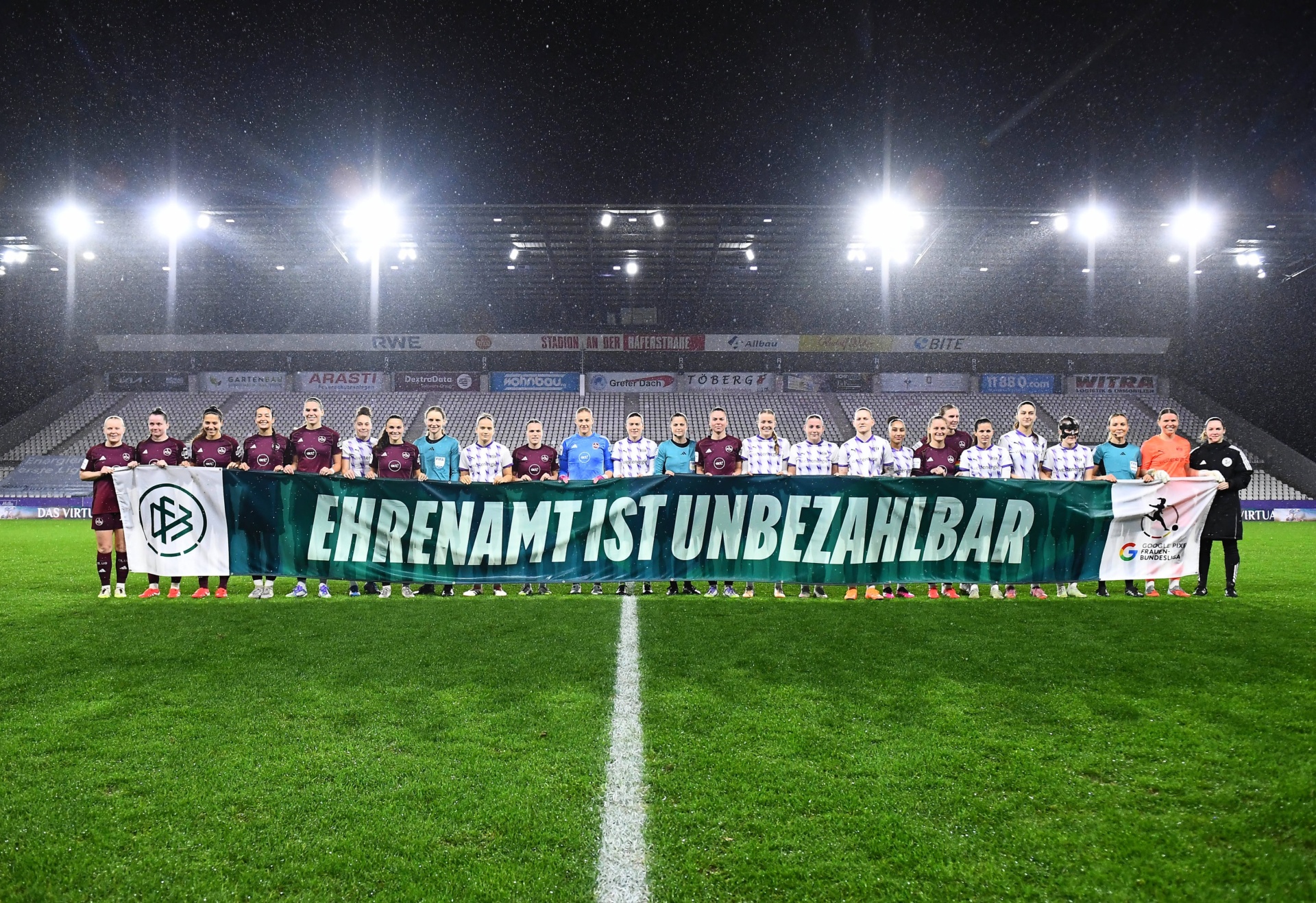 [{"clang_id":1,"value":"Fußballmannschaft in dunkelroten und weißen Trikots steht auf grünem Rasen vor einer Stadiontribüne. Sie halten eine grüne Banner mit weißem Schriftzug: \"EHRENAMT IST UNBEZAHLBAR\". Es regnet."}]