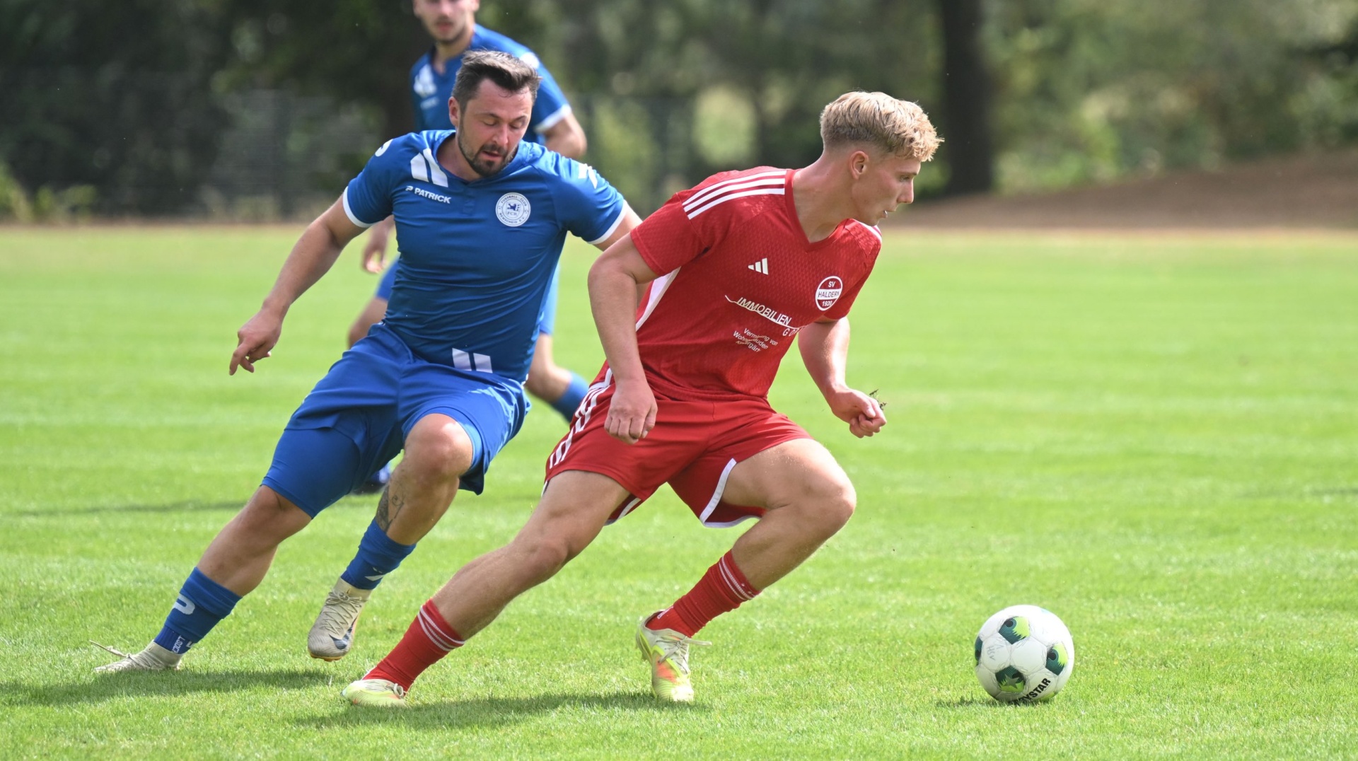 Zwei Fußballspieler, einer rot und einer blau gekleidet, laufen auf einem Rasenplatz.
