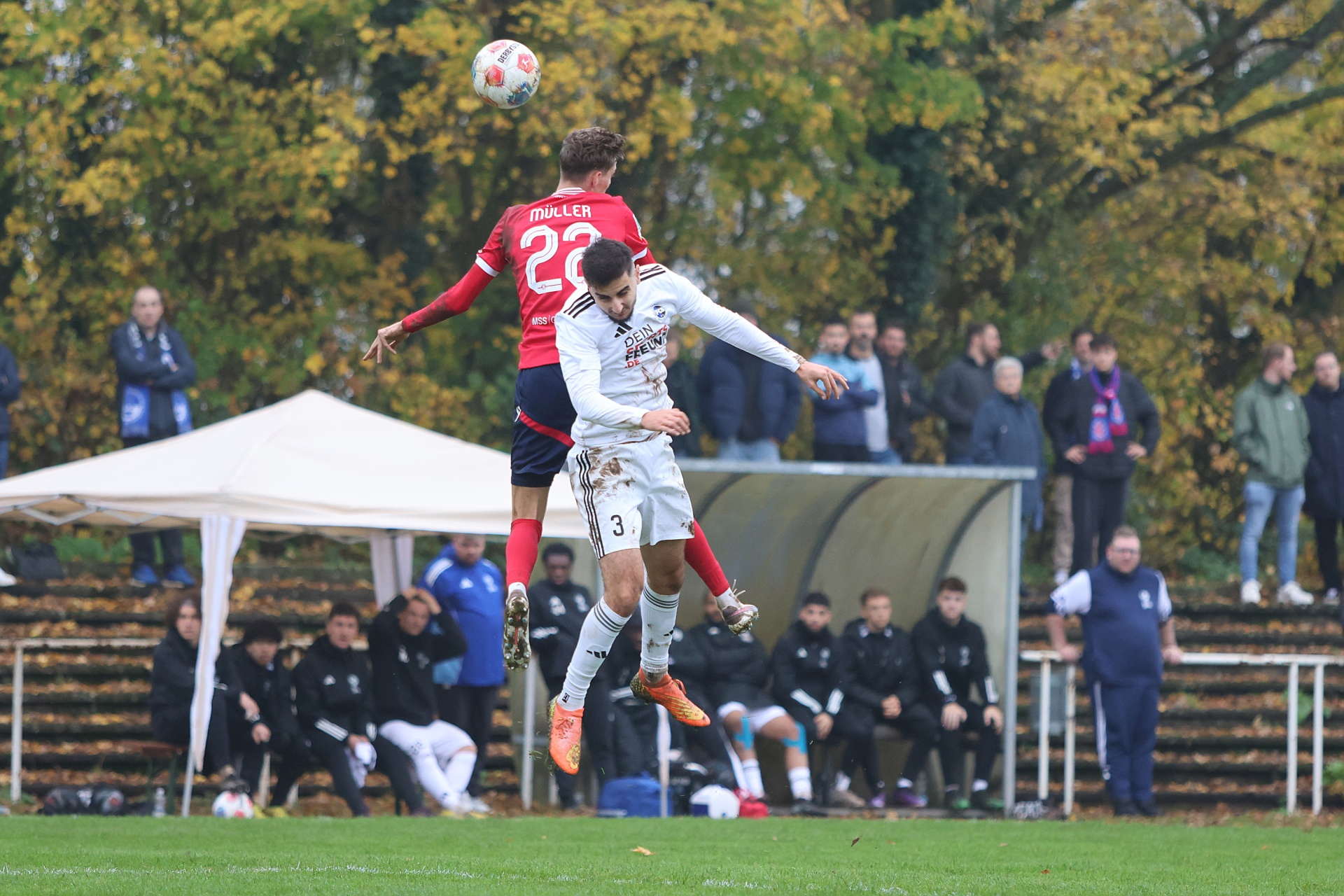 [{"clang_id":1,"value":"Zwei Fußballspieler springen im Spiel um den Ball. Im Hintergrund Zuschauer und Bäume mit herbstlichem Laub. Rasenplatz."}]