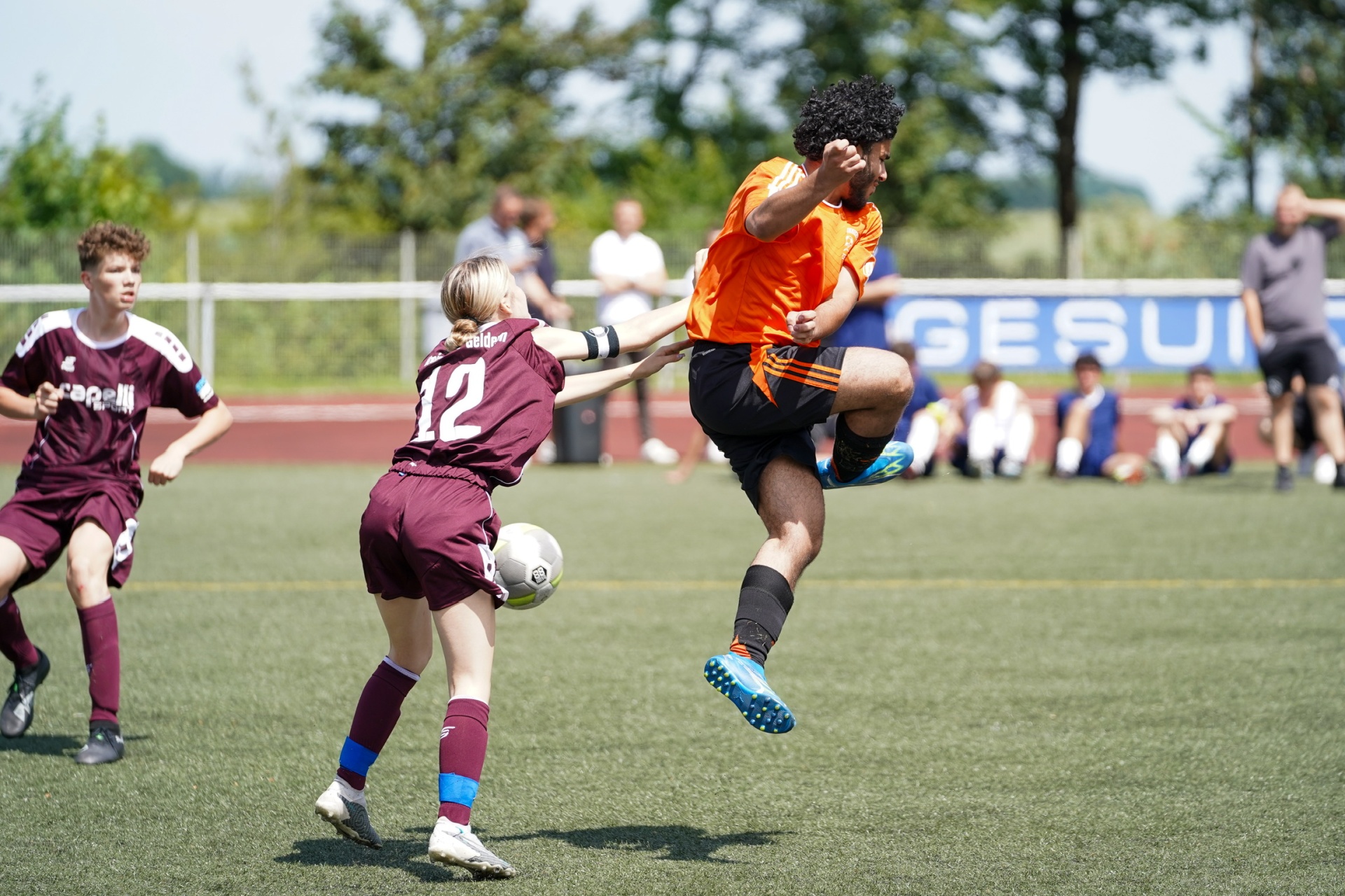 [{"clang_id":1,"value":"Fußballspiel: Spieler in orange und dunklen Trikots kämpfen um den Ball auf grünem Kunstrasen. Im Hintergrund Zuschauer und Bäume."}]