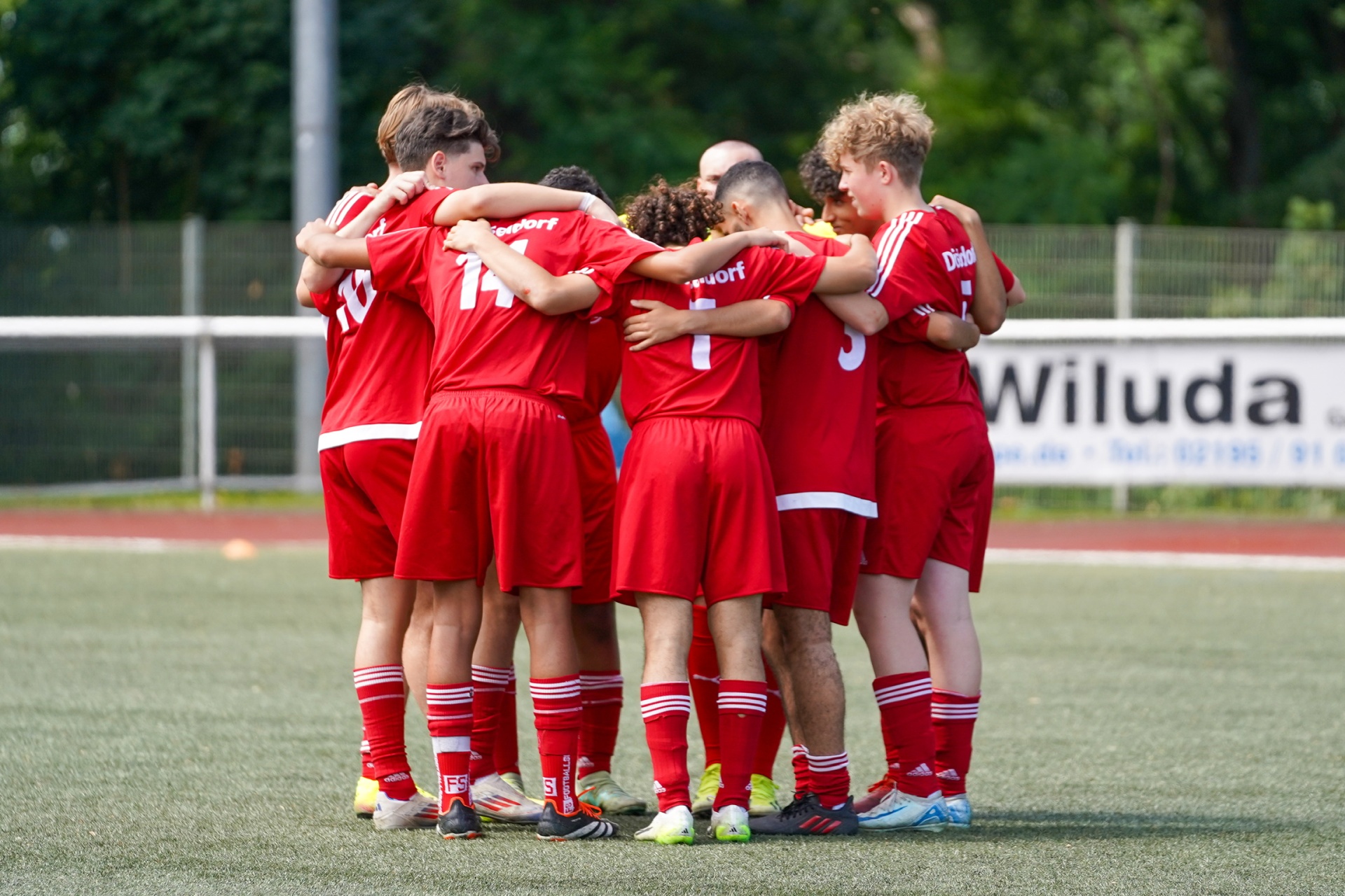 [{"clang_id":1,"value":"Fußballmannschaft in roten Trikots steht im Kreis und umarmt sich auf einem grünen Fußballfeld. Im Hintergrund eine Zuschauertribüne und Bäume."}]