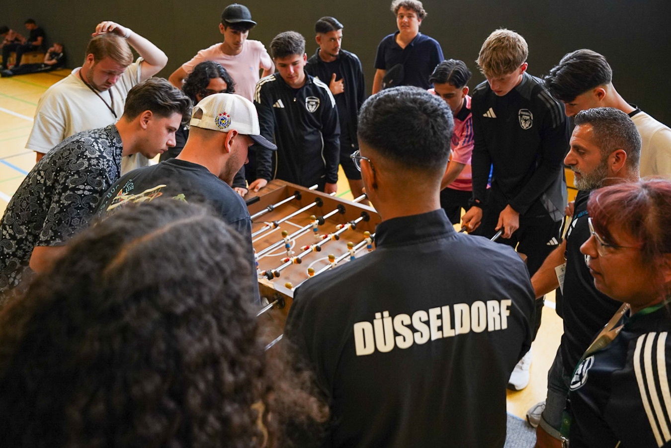 [{"clang_id":1,"value":"Gruppe von Männern um einen Tischfußballtisch in einer Sporthalle. Einige tragen Trikots mit der Aufschrift \"DÜSSELDORF\"."}]