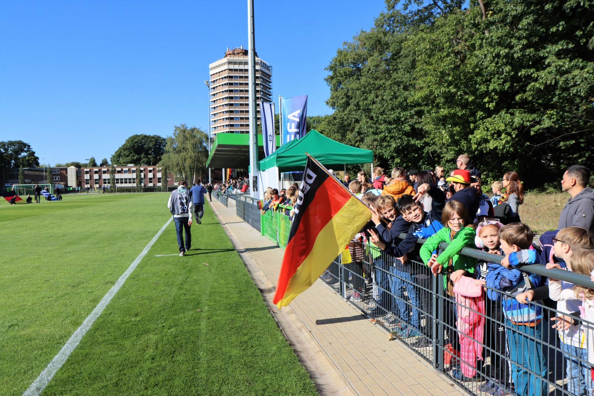 [{"clang_id":1,"value":"Fußballfeld mit Zuschauern an der Seitenlinie. Im Hintergrund Gebäude und Bäume unter blauem Himmel. Deutsche Flagge und Zelt sichtbar."}]