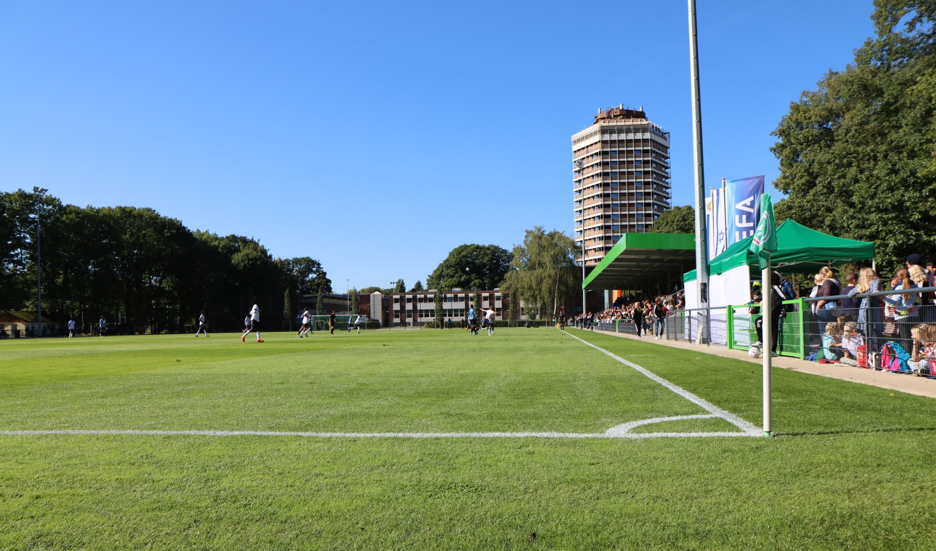 [{"clang_id":1,"value":"Fußballfeld mit Spielern bei sonnigem Wetter. Im Hintergrund Gebäude und Bäume, sowie ein kleines Zelt."}]