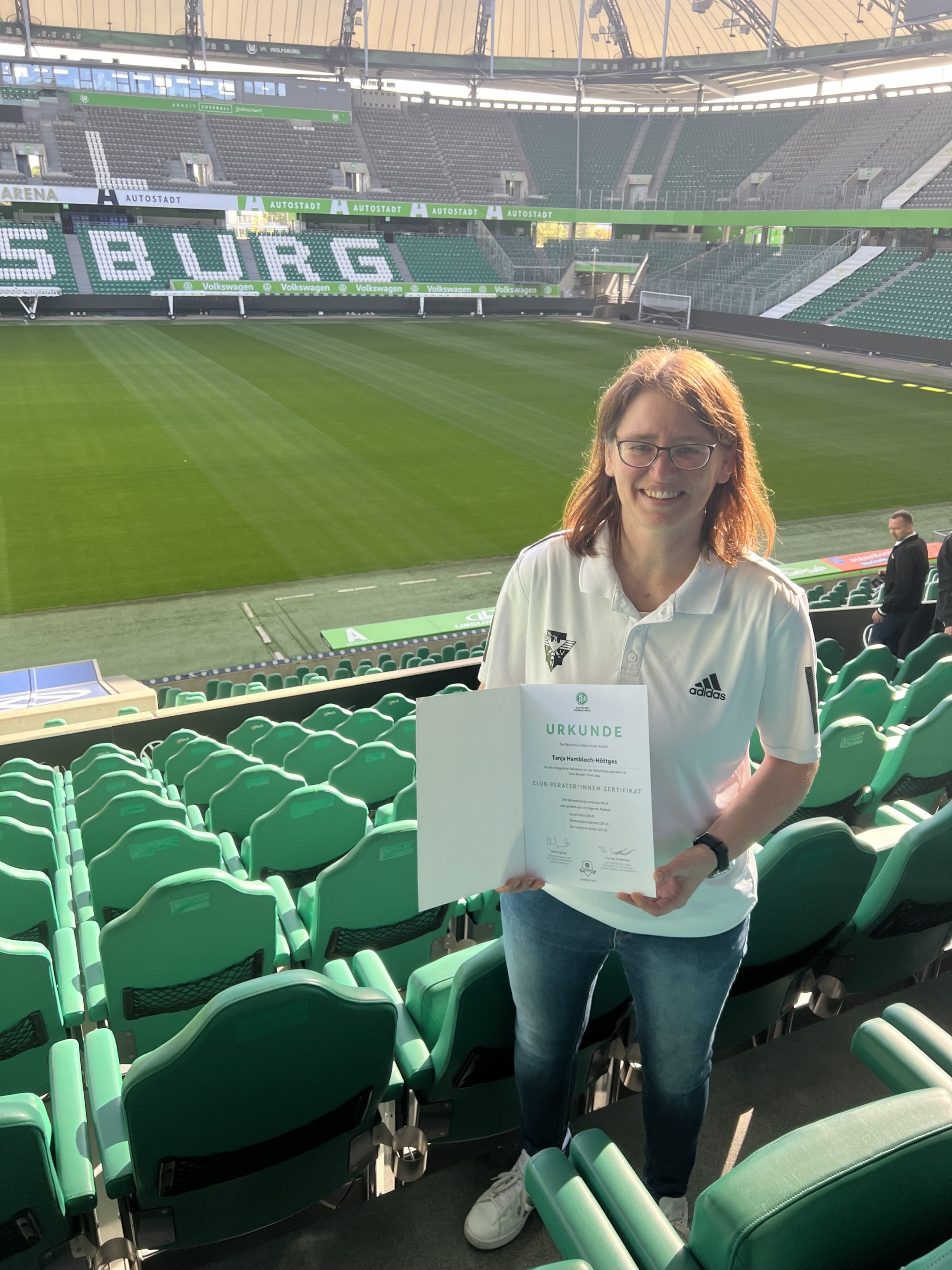 [{"clang_id":1,"value":"Frau mit Urkunde im Fußballstadion. Sie trägt ein weißes T-Shirt mit Logo und steht vor grünen Sitzreihen, Blick auf Rasen und Zuschauerränge."}]