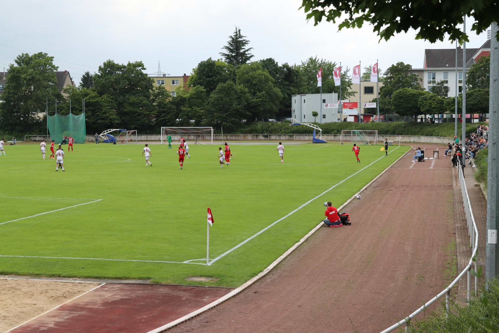 [{"clang_id":1,"value":"Fußballspiel auf grünem Rasenplatz. Spieler in roten und weißen Trikots. Zuschauerränge rechts, Bäume und Gebäude im Hintergrund. Roter Laufbahnrand."}]