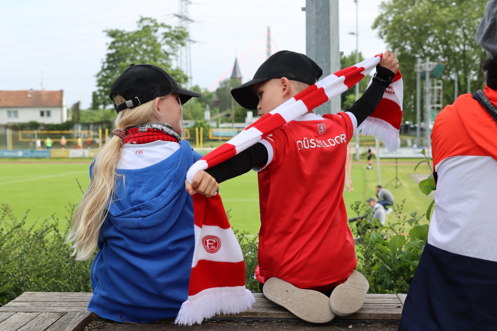 [{"clang_id":1,"value":"Drei Kinder stehen auf einer Mauer. Zwei tragen rot-weiße Fortuna Düsseldorf Schals und Trikots. Links trägt ein Kind eine blaue Jacke und eine Kappe. Im Hintergrund ein Fußballfeld."}]