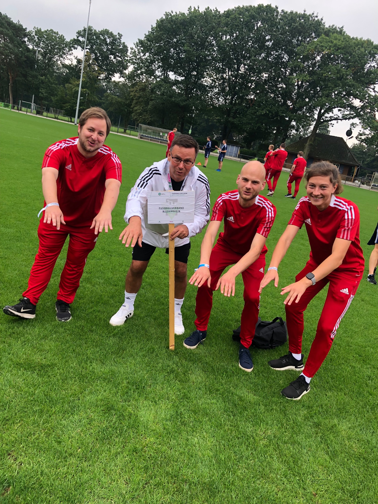 [{"clang_id":1,"value":"Fünf Männer in roten Trainingsanzügen knien auf einem Fußballfeld um einen Stock, der im Boden steckt. Im Hintergrund sind weitere Personen und Bäume zu sehen."}]